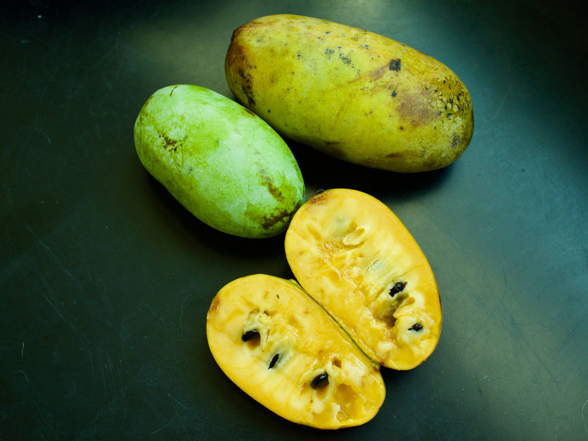 Fruit 桌布 壁紙 — 三個泡泡果放在深色表面上，一個完整綠色，一個完整黃色，一個切開露出奶油色果肉和黑色種子。