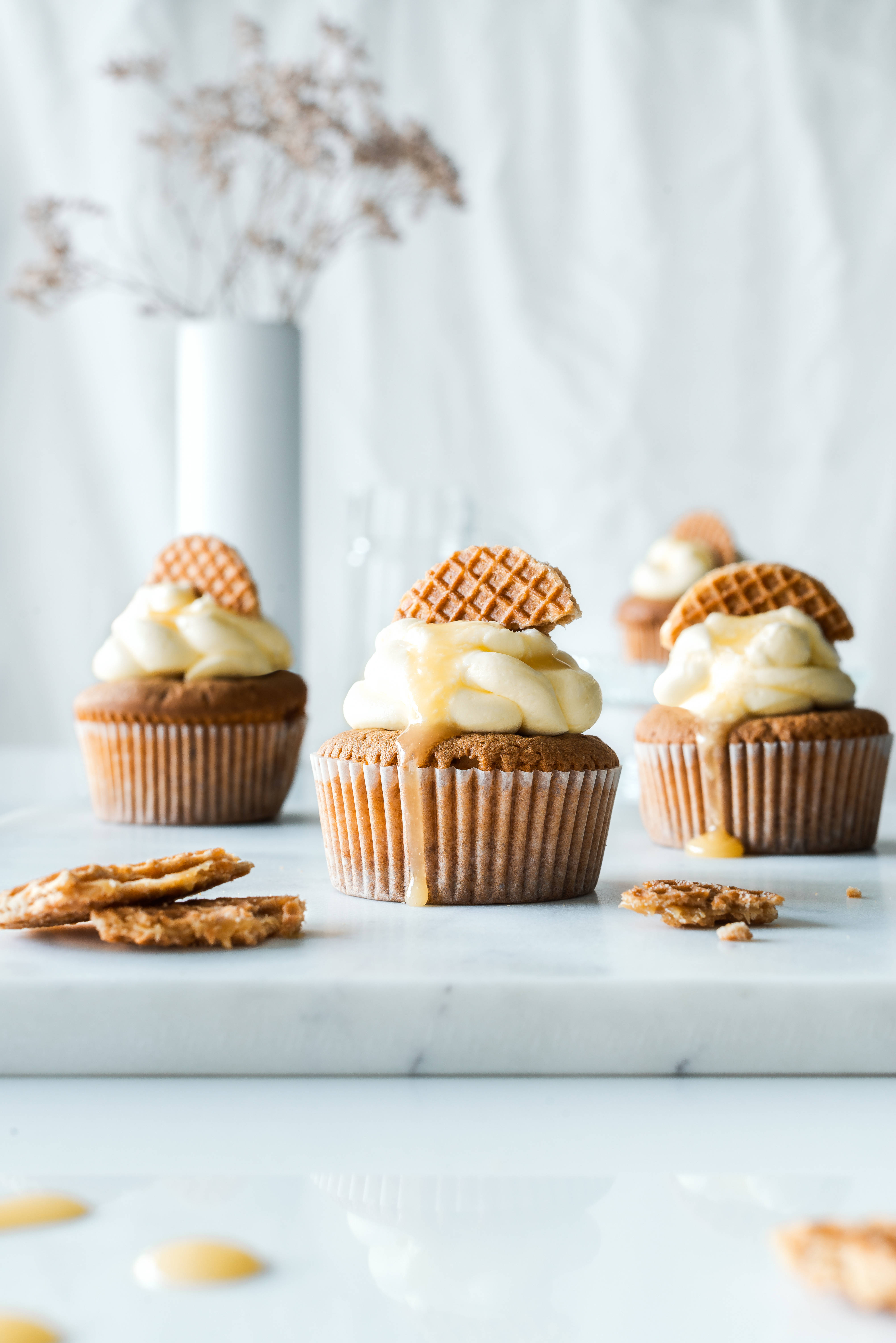 Cupcake 桌布 壁紙 — 三個淋上焦糖醬、頂部放有鬆餅餅乾的杯子蛋糕，置於大理石表面上，周圍散落著碎屑，背景有一瓶乾花。