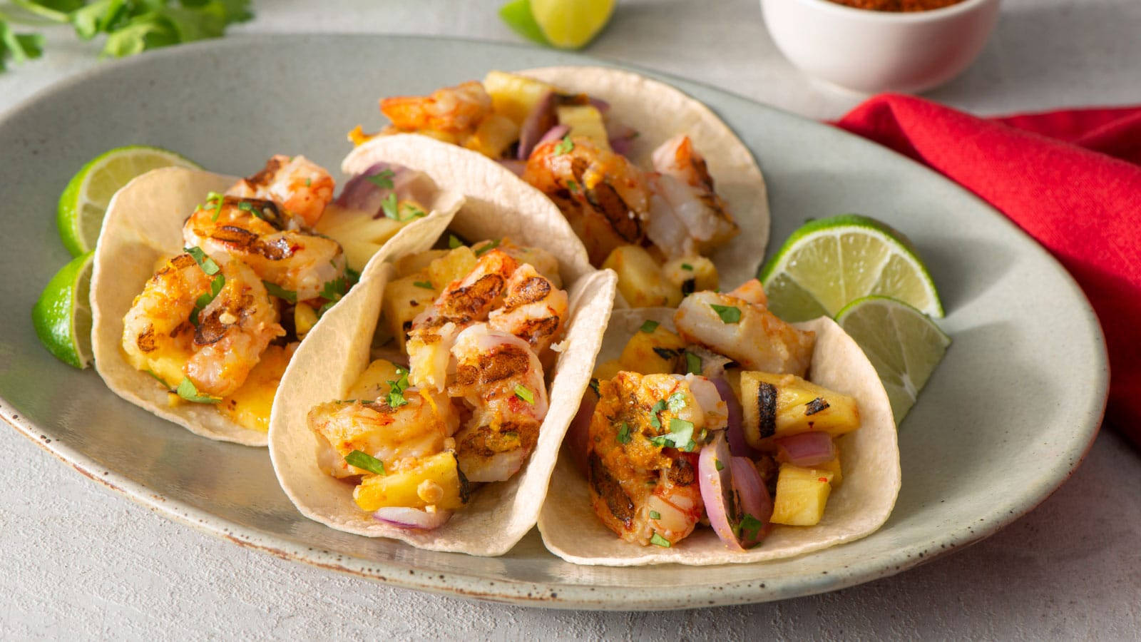 Tacos 桌布 壁紙 — 三個烤蝦玉米餅配鳳梨洋蔥和香菜，盛在灰色盤子上，附有萊姆角和紅色餐巾。