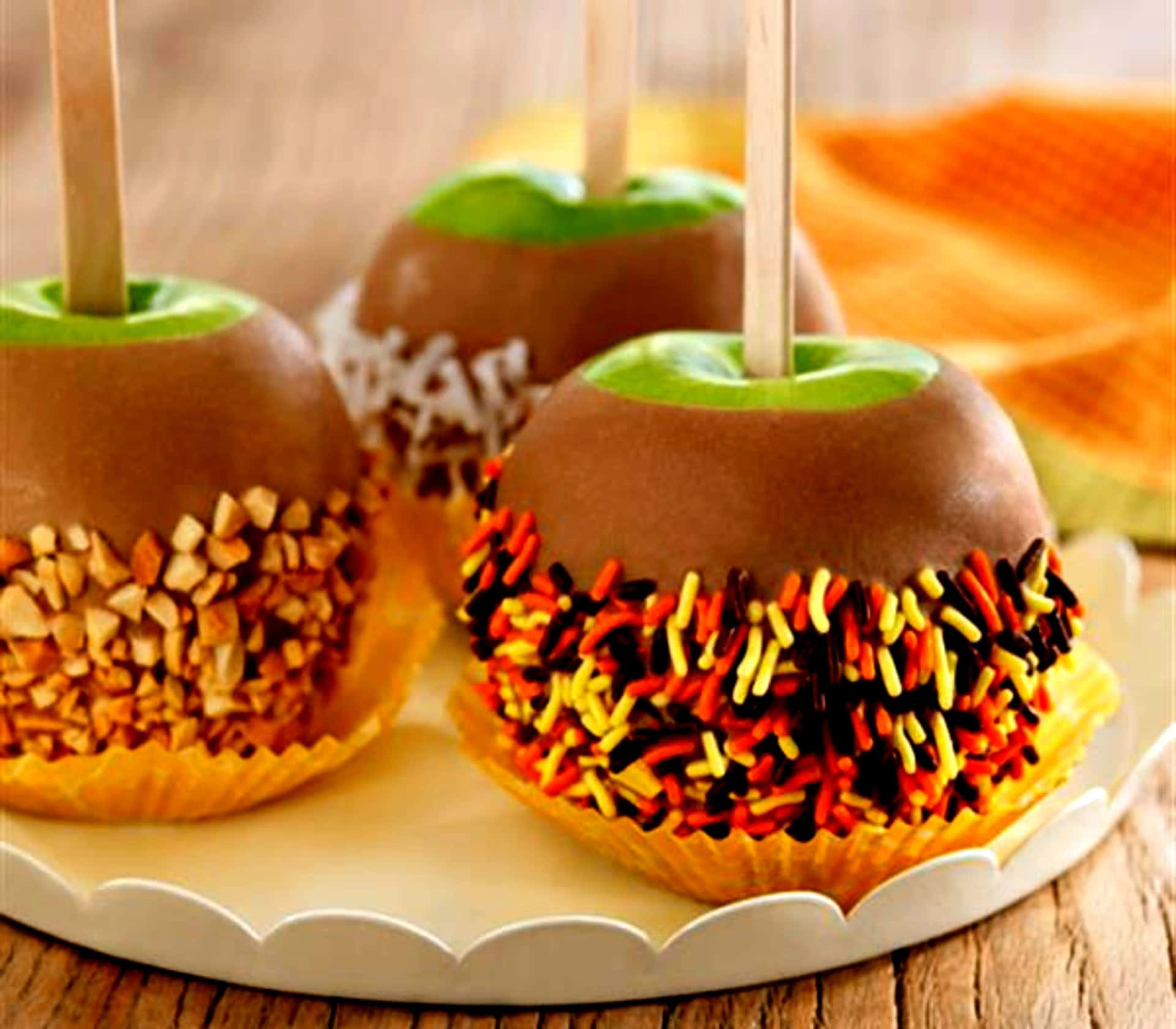 Caramel Apples 桌布 壁紙 — 三個焦糖蘋果，外層裹上巧克力，並以堅果或彩色糖粒裝飾，放在一個白色扇貝邊盤子上。
