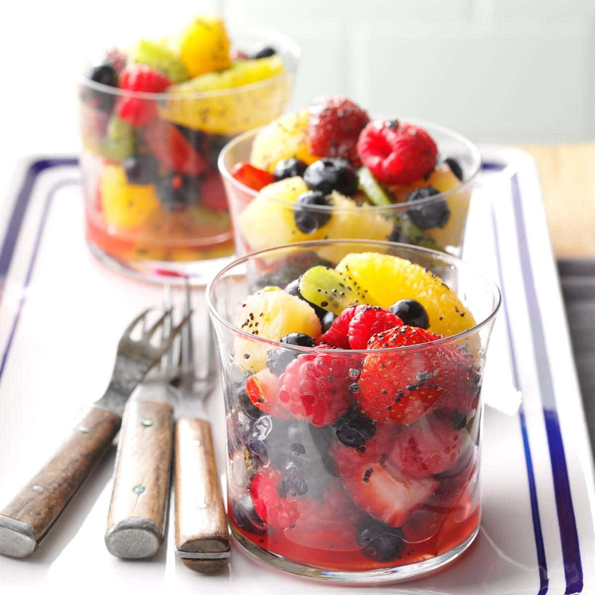 Fruit 桌布 壁紙 — 三個玻璃杯盛滿色彩繽紛的水果沙拉，包括草莓、覆盆子、藍莓、奇異果和鳳梨，放在白色托盤上，附有木柄叉子。