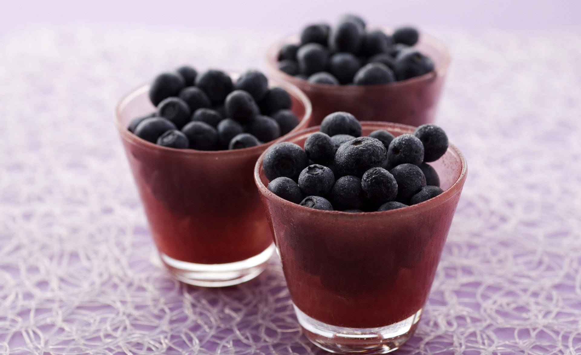 Blueberry 桌布 壁紙 — 三個玻璃杯裝滿鮮豔的藍莓和紅色液體，放在精緻的白色蕾絲桌布上，背景是柔和的紫色。