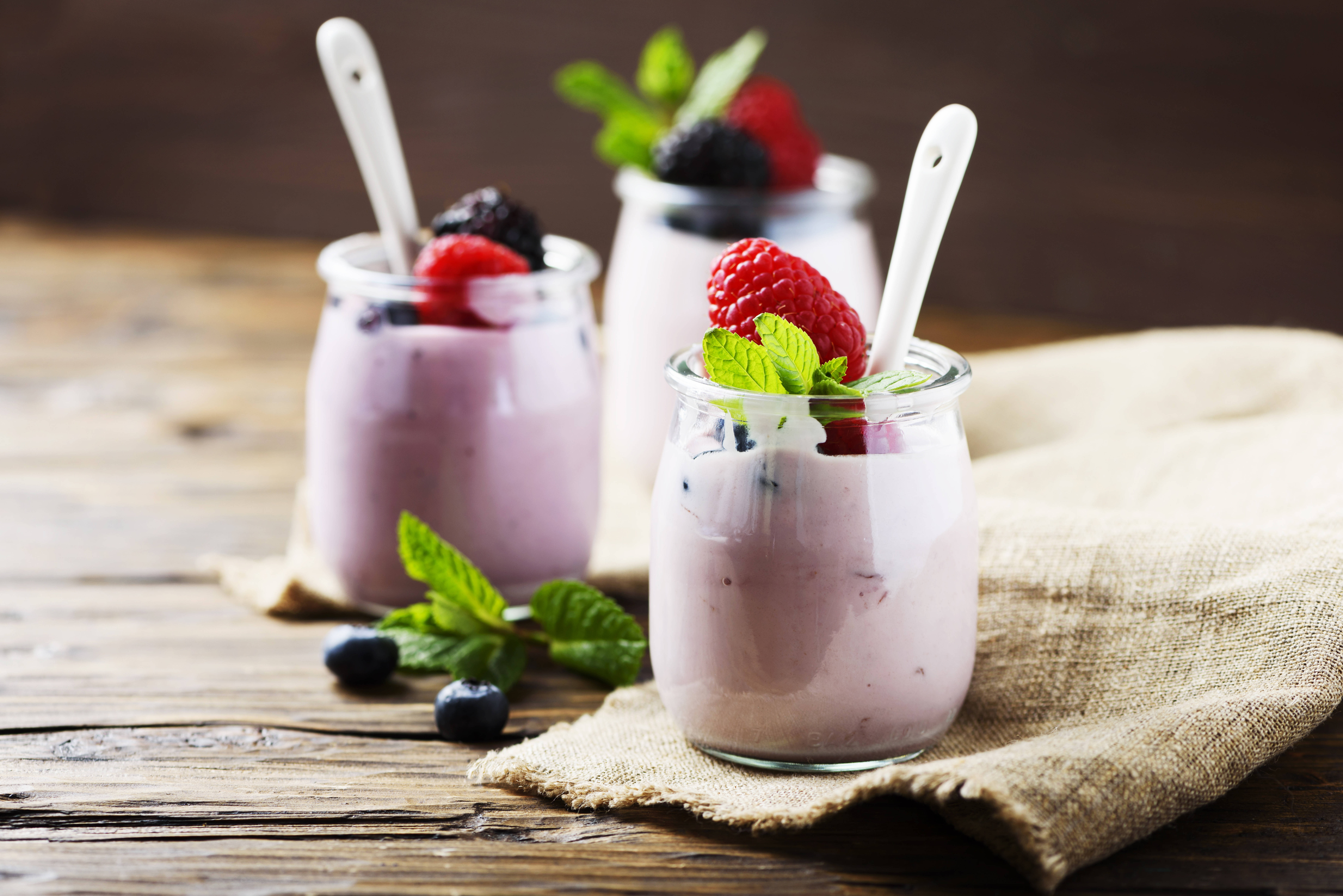 Yogurt 桌布 壁紙 — 三個玻璃罐裝滿粉紅色莓果優格，上面放上新鮮覆盆子、黑莓和薄荷葉，放在質樸的木桌上。