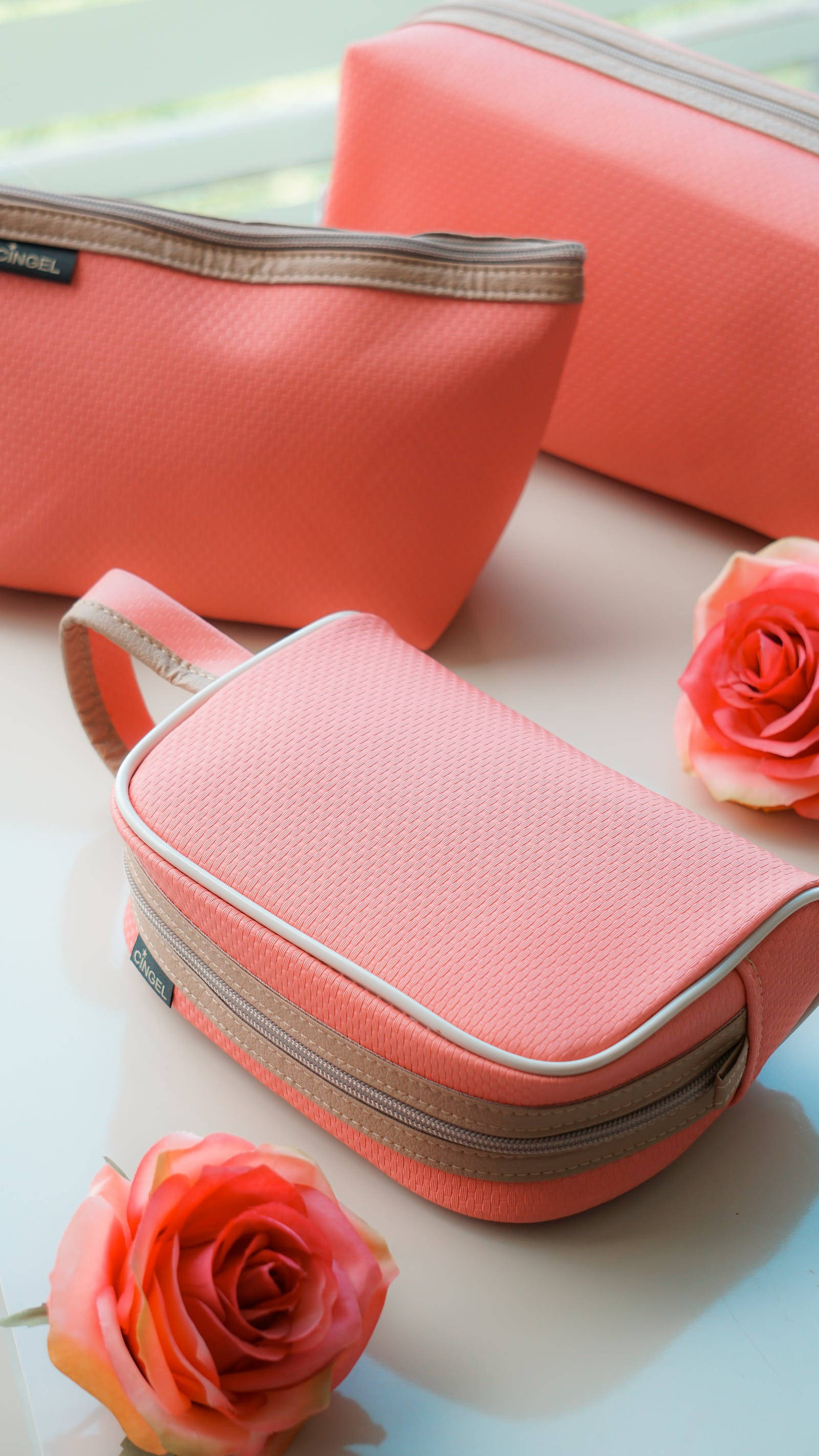 Rose iPhone 桌布 壁紙 — 三個珊瑚粉紅色紋理化化妝包配米色拉鍊，放在白色表面上，旁邊有兩朵盛開的珊瑚玫瑰。