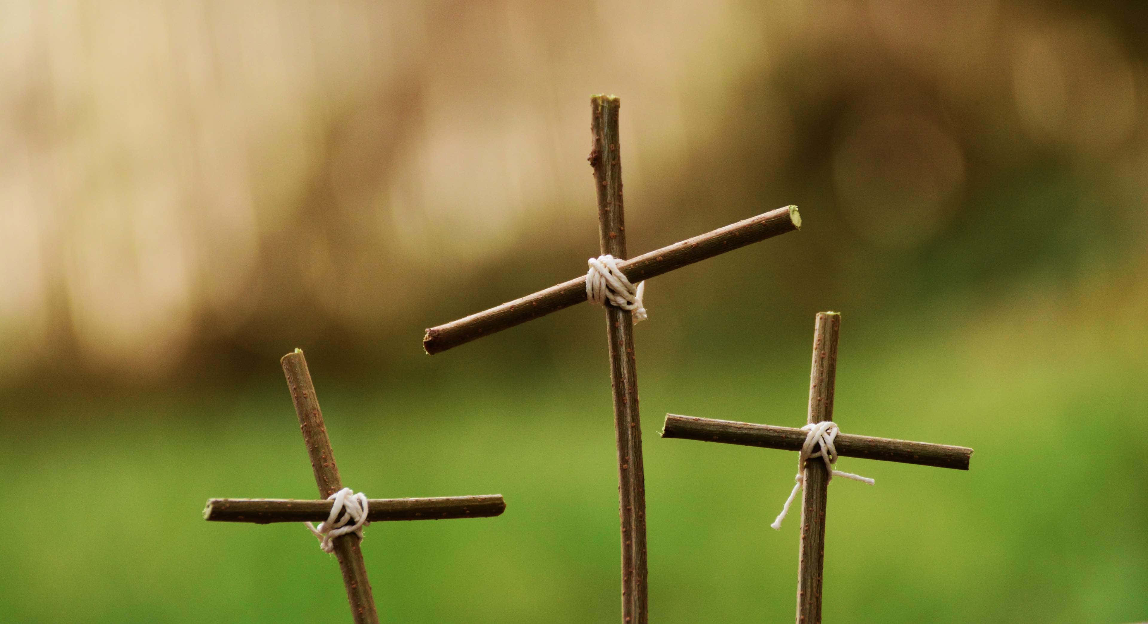 Jesus Crucifixion 桌布 壁紙 — 三個用白色繩子綁著的鄉村風格樹枝十字架，在模糊的綠色和棕色自然背景中以柔焦呈現。
