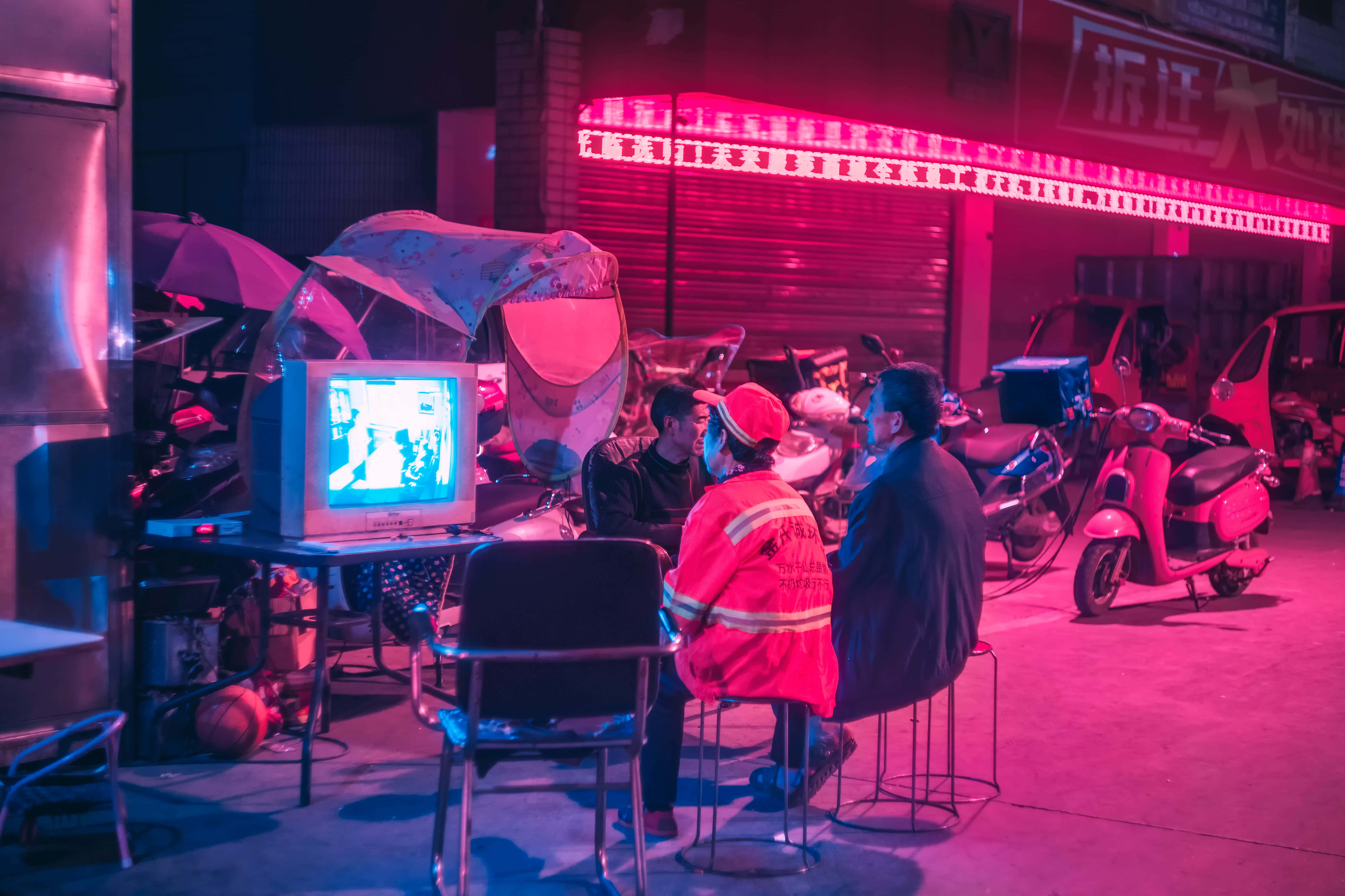 watching tv 桌布 壁紙 — 三個男人圍著一部發出藍光的舊CRT電視，身處夜晚霓虹燈閃爍的小巷，周圍是機車和已關門的商店。