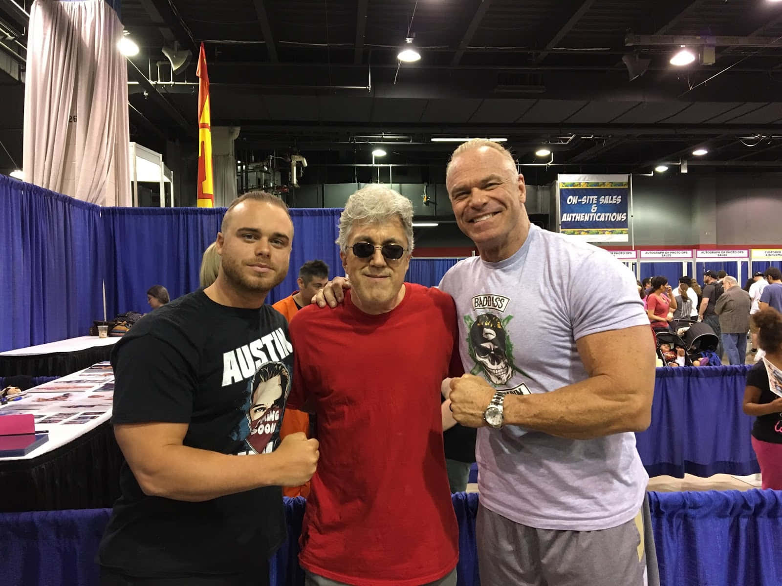 WWE 桌布 壁紙 — 三個男人在一個熙熙攘攘的會議廳裡一起擺姿勢，廳內有藍色桌布覆蓋的桌子和頭頂燈光。