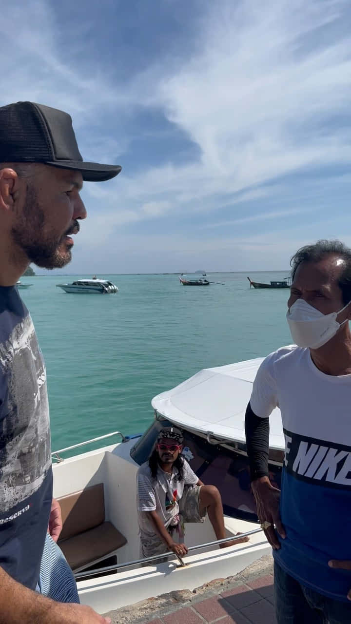 MMA 桌布 壁紙 — 三個男人在一個熱帶碼頭的白船上，周圍有藍綠色海水和遠處的船，天空局部多雲。