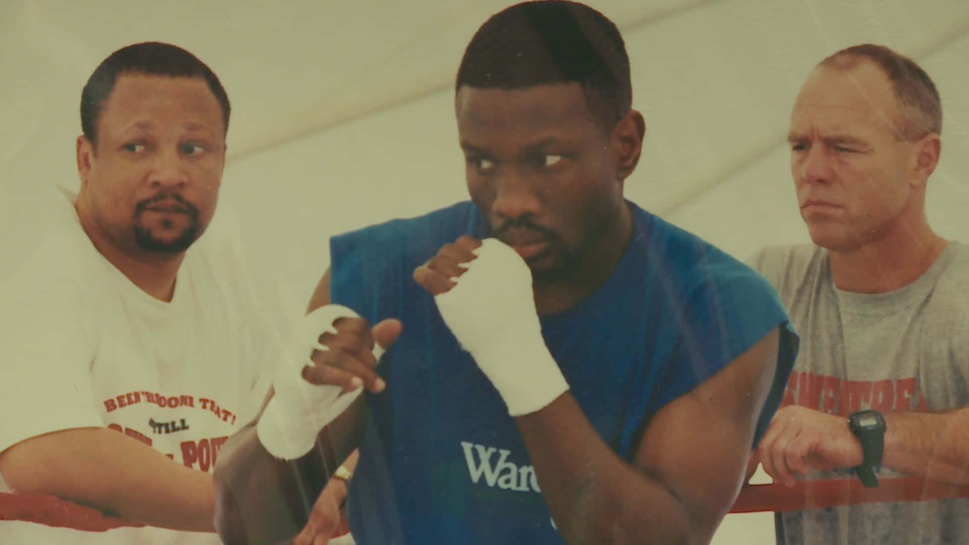 Boxing 桌布 壁紙 — 三個男人在拳擊館，其中一名身穿藍色背心、雙手纏繃帶的拳手處於專注姿勢。