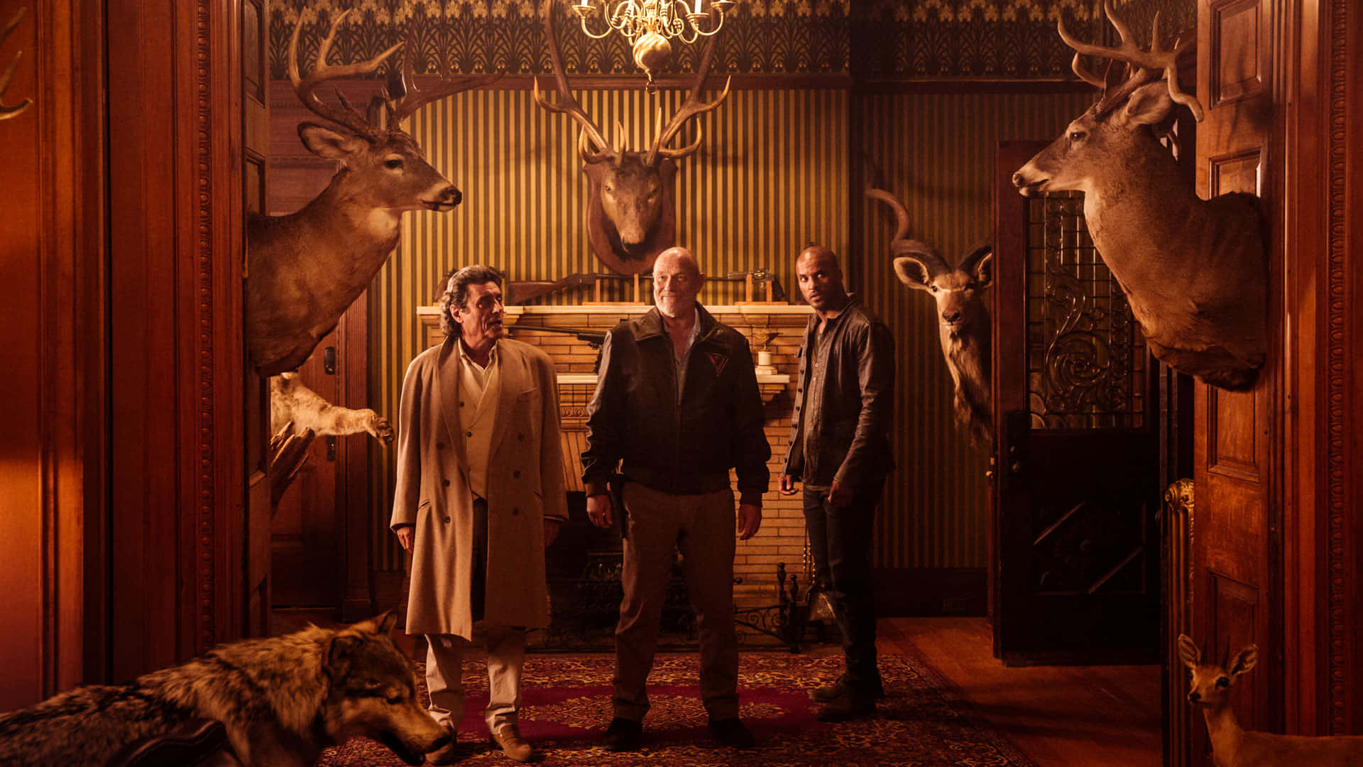 American Gods 桌布 壁紙 — 三個男人站在一個燈光溫暖、木質鑲板的房間裡，房間裝飾著掛起的鹿頭和一隻地上的狼標本。