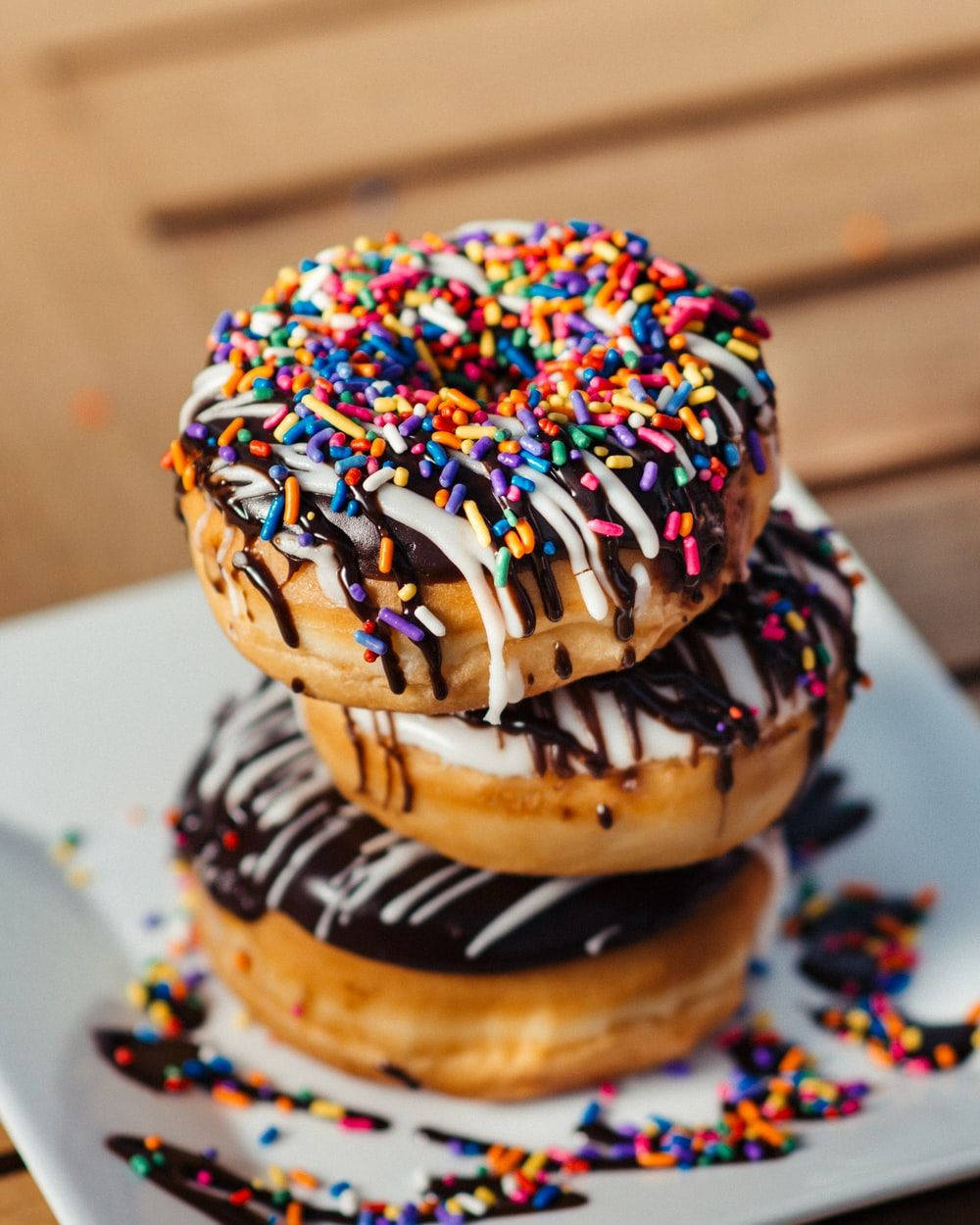 donuts 桌布 壁紙 — 三個疊起的巧克力糖霜甜甜圈，飾有彩虹糖針和白色糖漿，放在白色盤子上。
