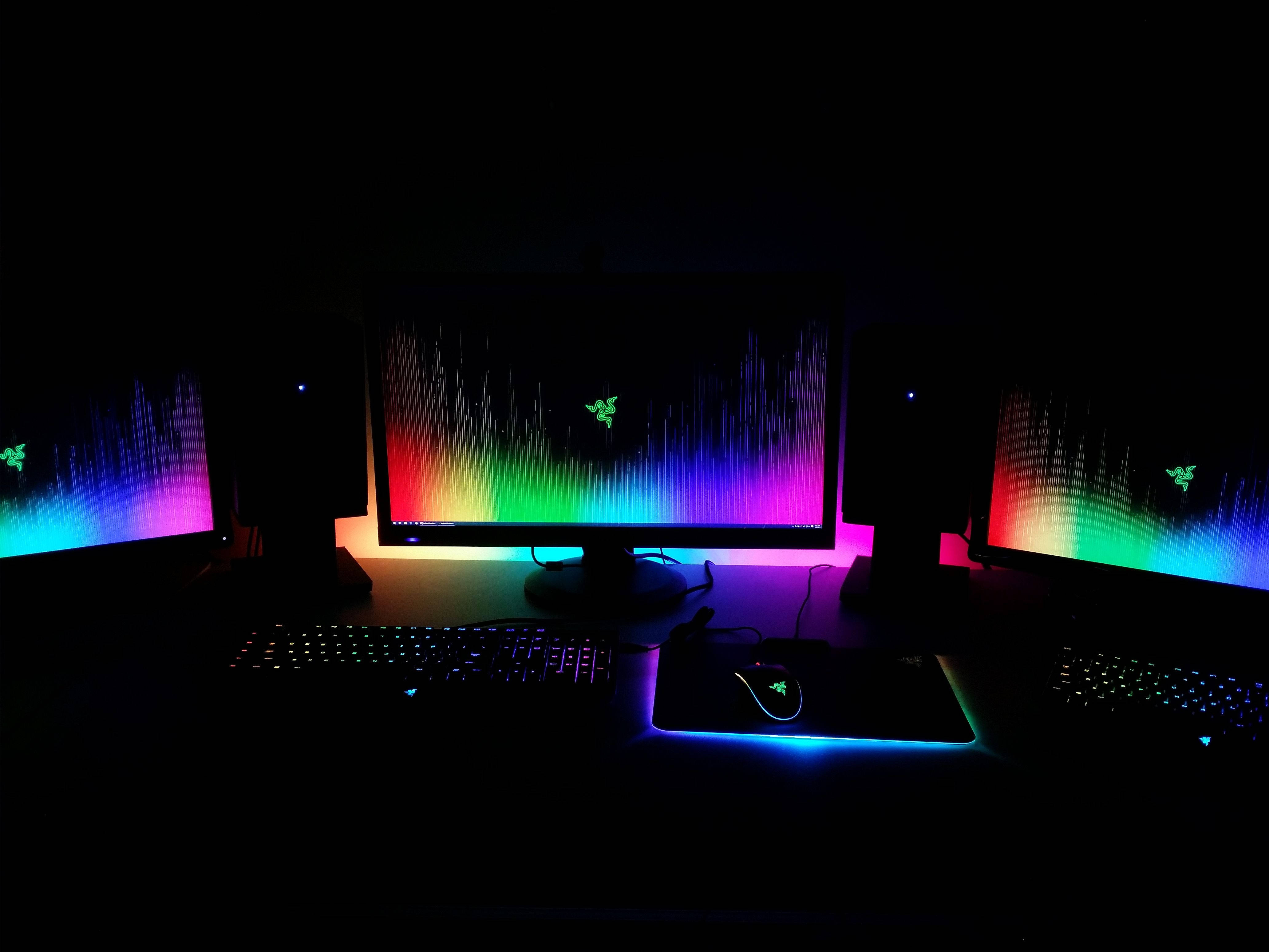 room 桌布 壁紙 — 三個發光的Razer顯示器在黑暗的遊戲設置中顯示充滿活力的彩虹波浪桌布，並配有發光的外圍設備。