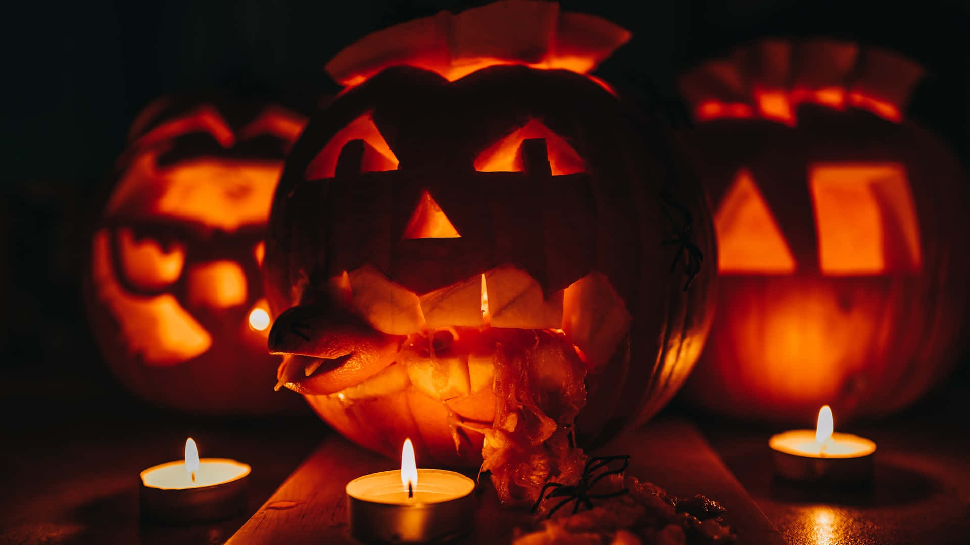 Halloween Candles 桌布 壁紙 — 三個發光的傑克南瓜燈，帶有雕刻的臉和蠟燭，在黑暗的環境中投射出詭異的橙色光芒。