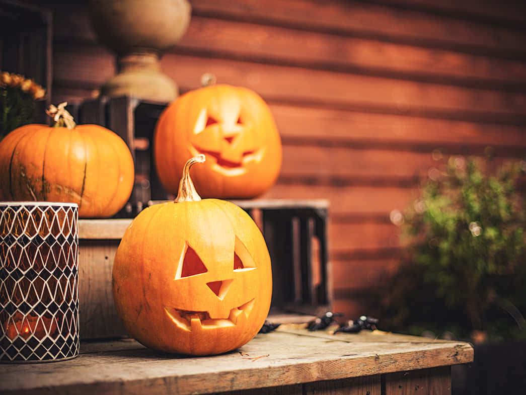 Jack O Lantern 桌布 壁紙 — 三個發光的傑克南瓜燈，帶有雕刻的臉孔，放在一個質樸的木桌上，旁邊有一個金屬網架內的點燃蠟燭。
