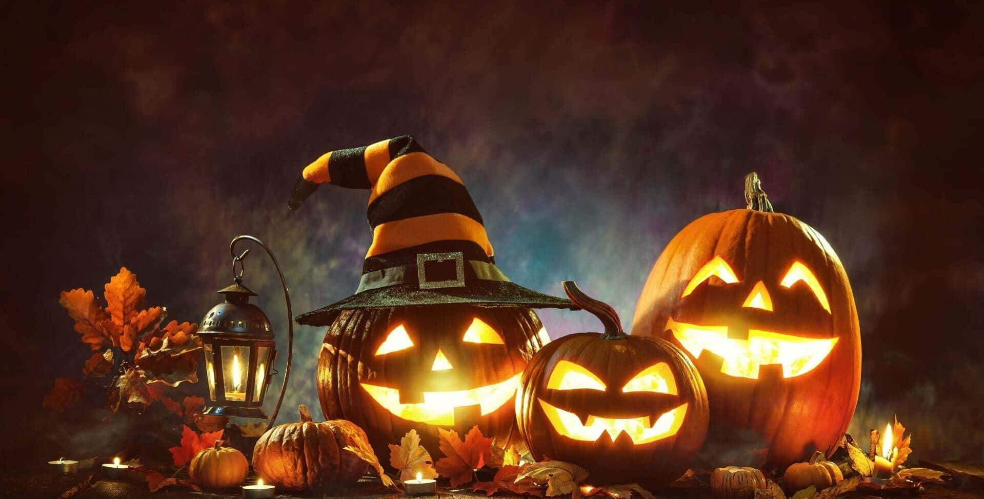 Jack O Lantern 桌布 壁紙 — 三個發光的南瓜燈，配上女巫帽和燈籠，在黑暗的天空下將詭異的光芒投射在秋葉上。