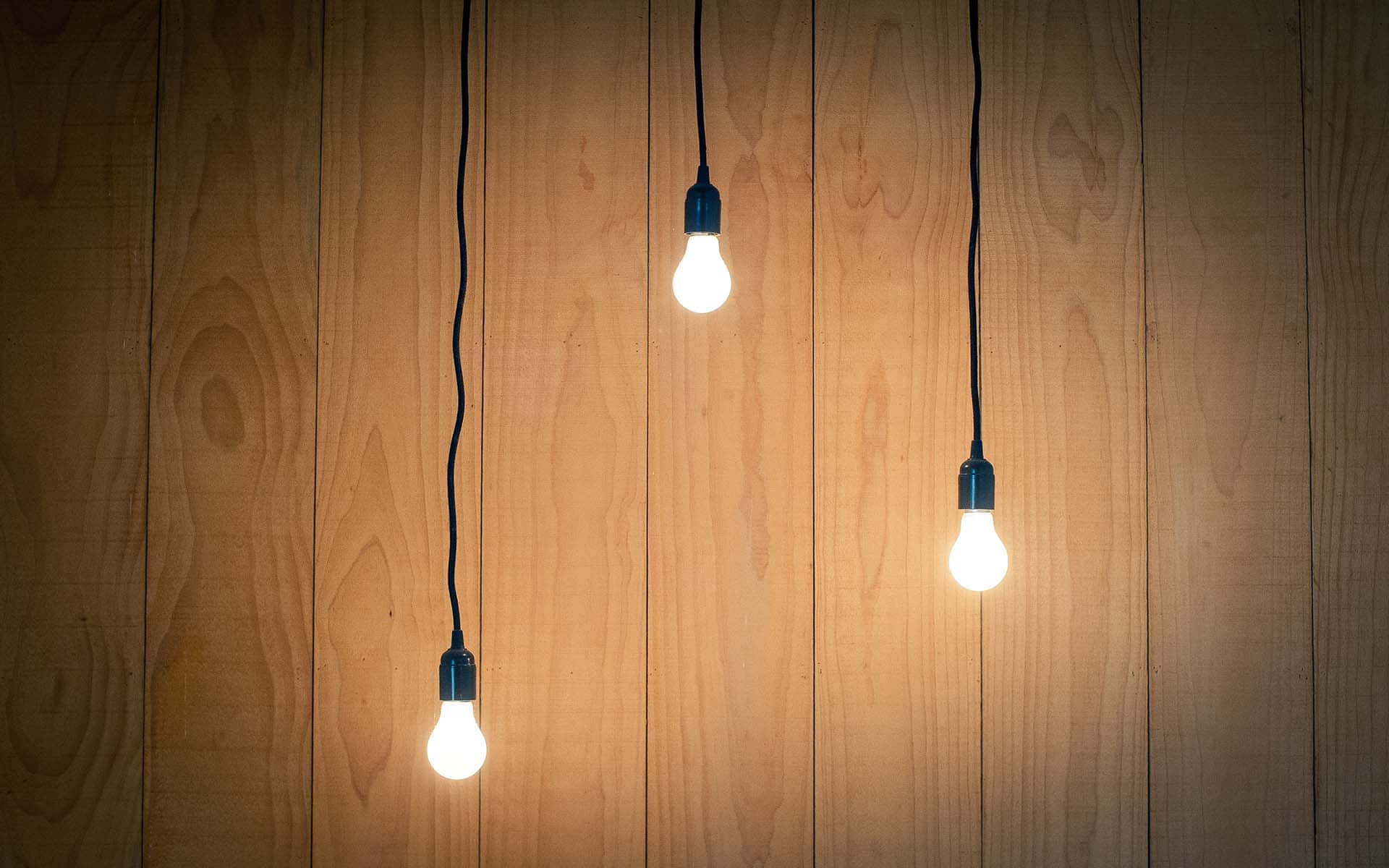 Light 桌布 壁紙 — 三個發光的燈泡用黑色電線掛在溫暖的木板上，投射出柔和的金色光芒。
