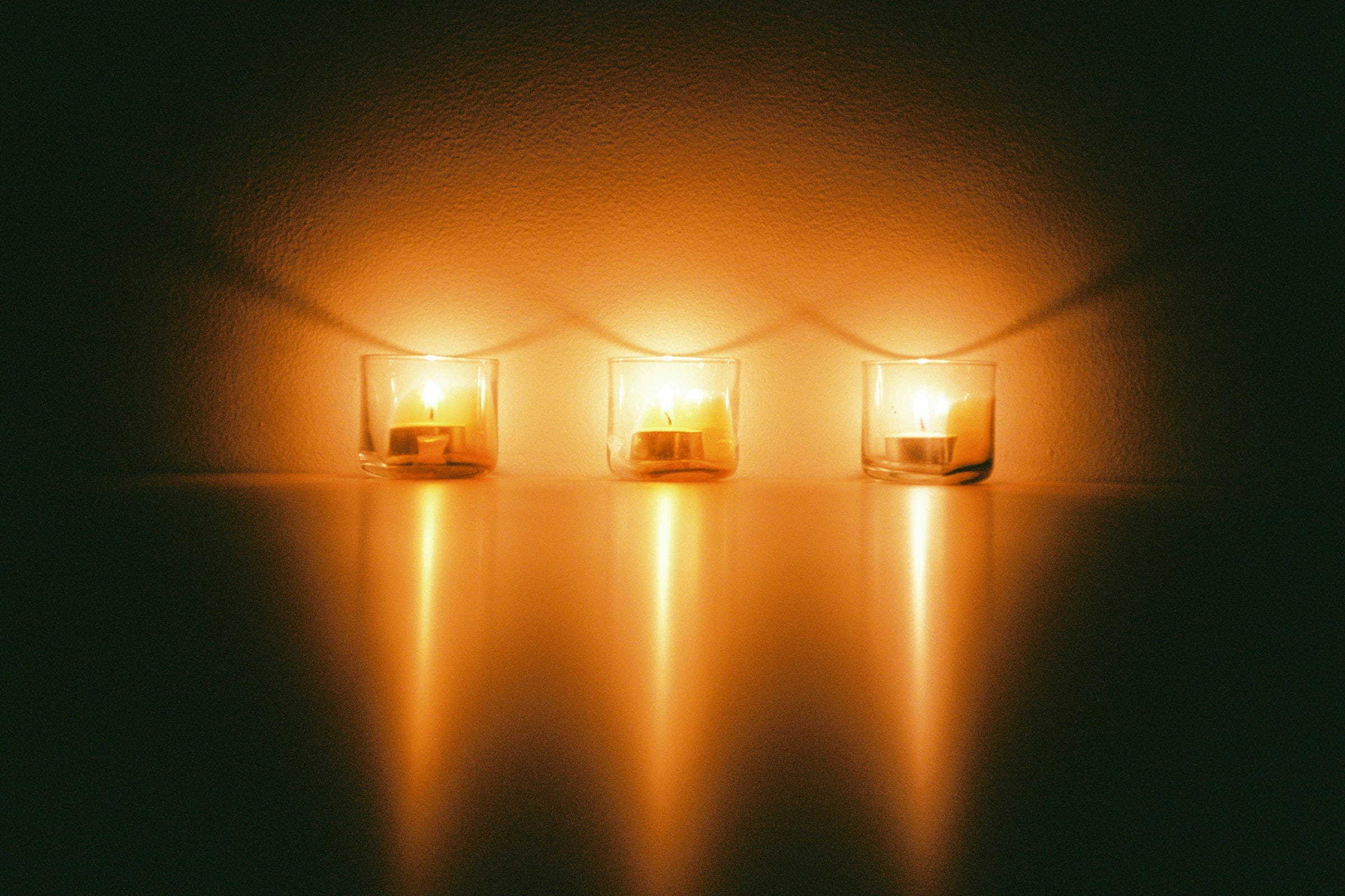 Remembrance 桌布 壁紙 — 三個發光的茶燭在玻璃燭台中，在深色表面上投射出溫暖的金色倒影，背景為有紋理的牆壁。