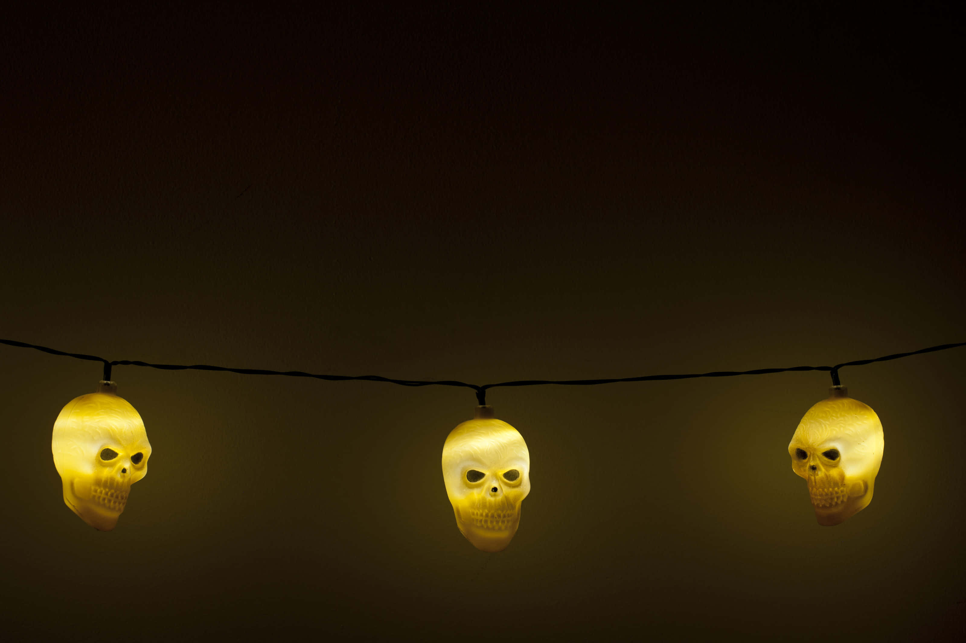 Spooky Halloween 桌布 壁紙 — 三個發光的黃色骷髏形燈泡掛在一條黑色繩子上，背景是深色漸變。