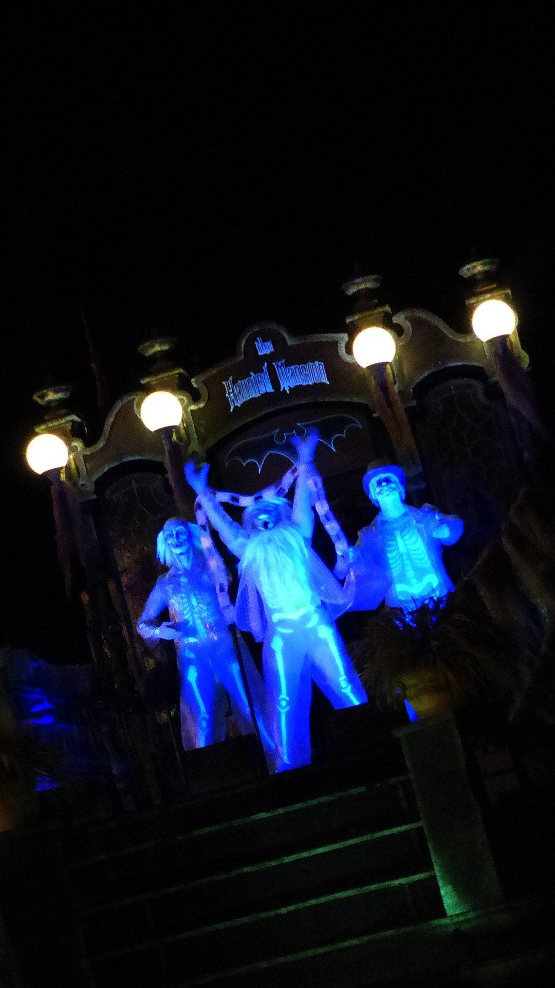 Scary Halloween iPhone 桌布 壁紙 — 三個發光藍色骷髏人物在夜間於鬼屋入口處的溫暖燈籠下戲劇性地擺姿勢。