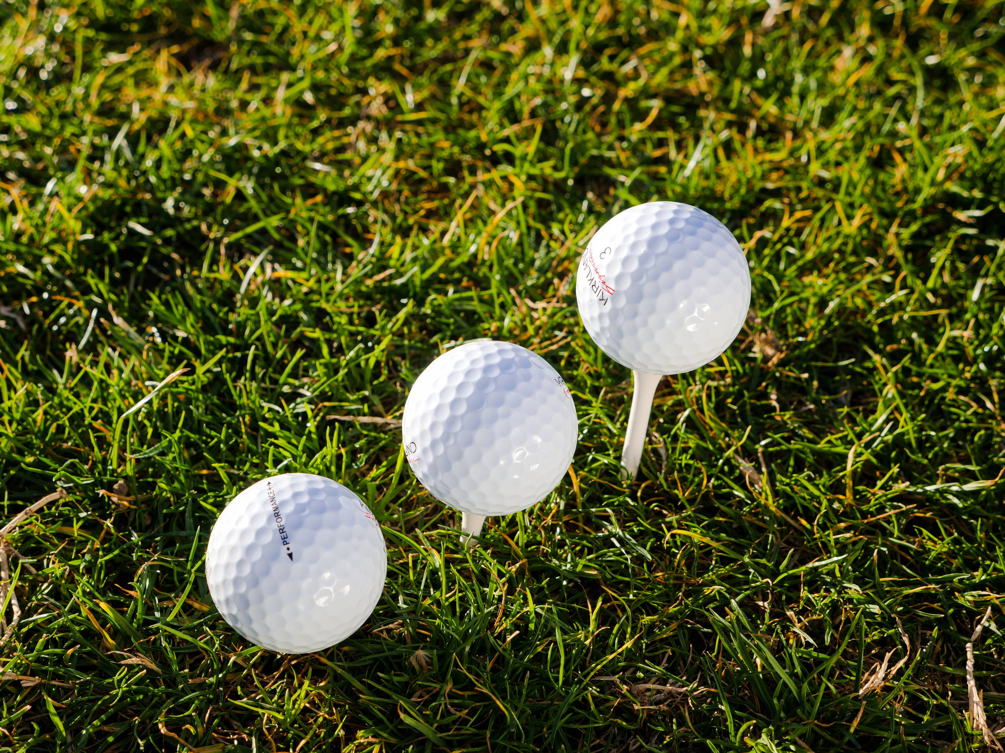 4K Golf 桌布 壁紙 — 三個白色高爾夫球在明亮的陽光下放在鮮綠色的草地上，其中一個放在球座上。