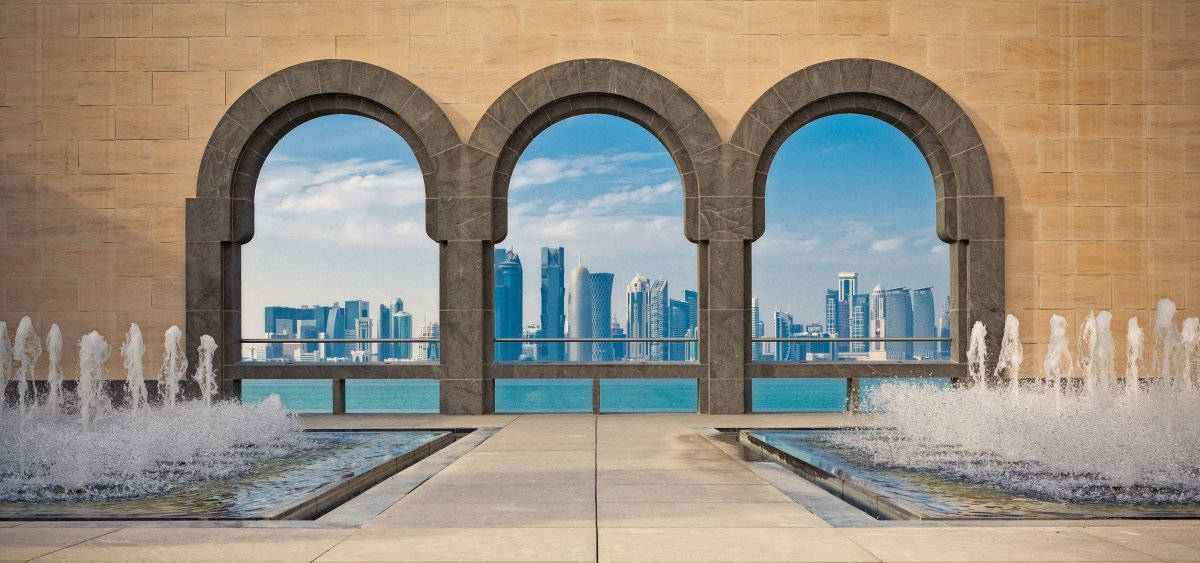 Qatar 桌布 壁紙 — 三個石拱門框住現代城市天際線，下方是碧綠海水，前景有噴泉噴灑。