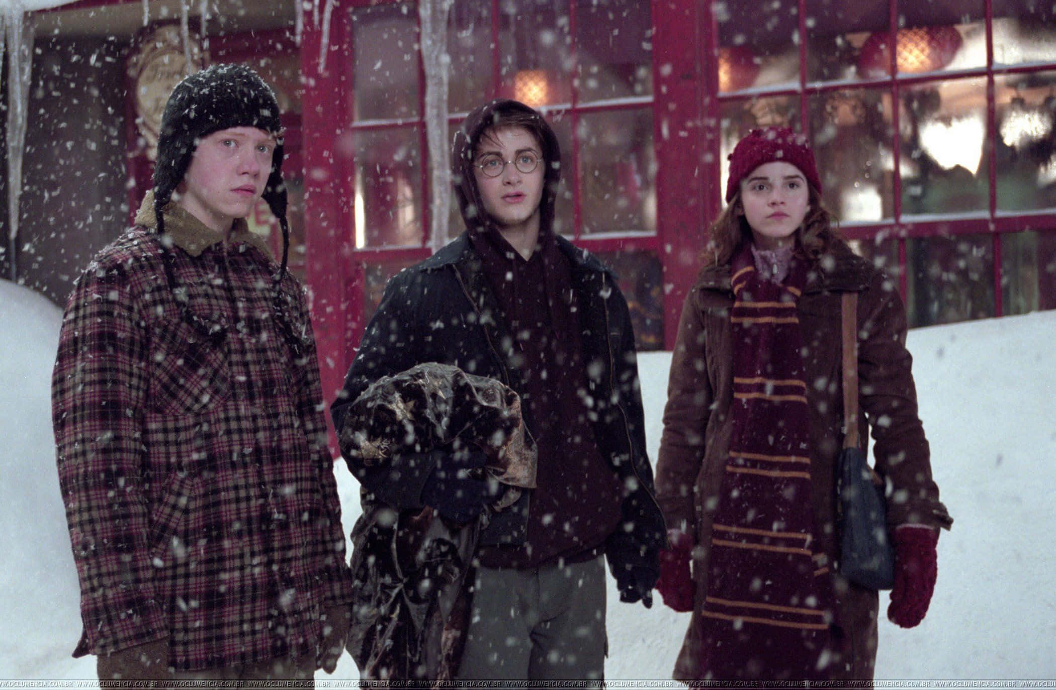 Golden Trio 桌布 壁紙 — 三個穿著冬季外套的年輕人站在飄雪中，外面是一座紅色框架的建築物，窗戶發著光。