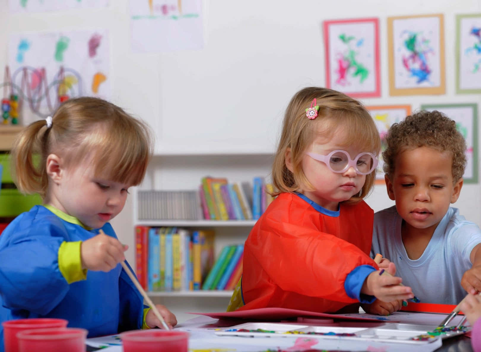 Learning 桌布 壁紙 — 三個穿著彩色罩衫的幼兒在充滿活力藝術品和書籍的桌子旁專心繪畫。
