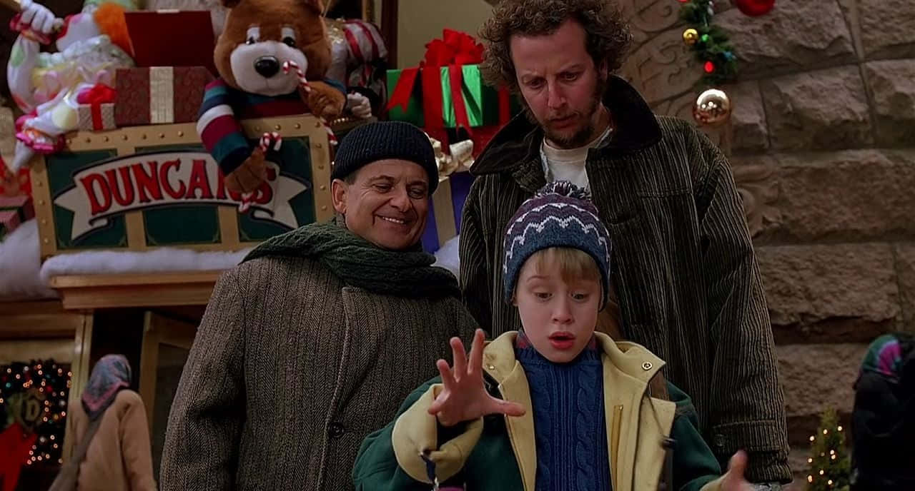Funny Home Alone 桌布 壁紙 — 三個穿著節日冬季外套和帽子的人物站在裝飾有節日燈光和禮物的Duncan's糖果車前。