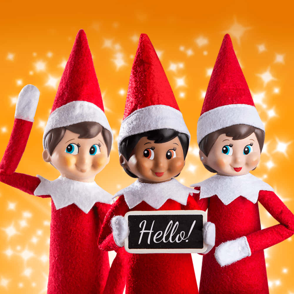 Elf On The Shelf 桌布 壁紙 — 三個穿著紅色服裝的快樂精靈公仔揮手並拿著一塊寫著「你好」的黑板，背景是閃爍的橙色。