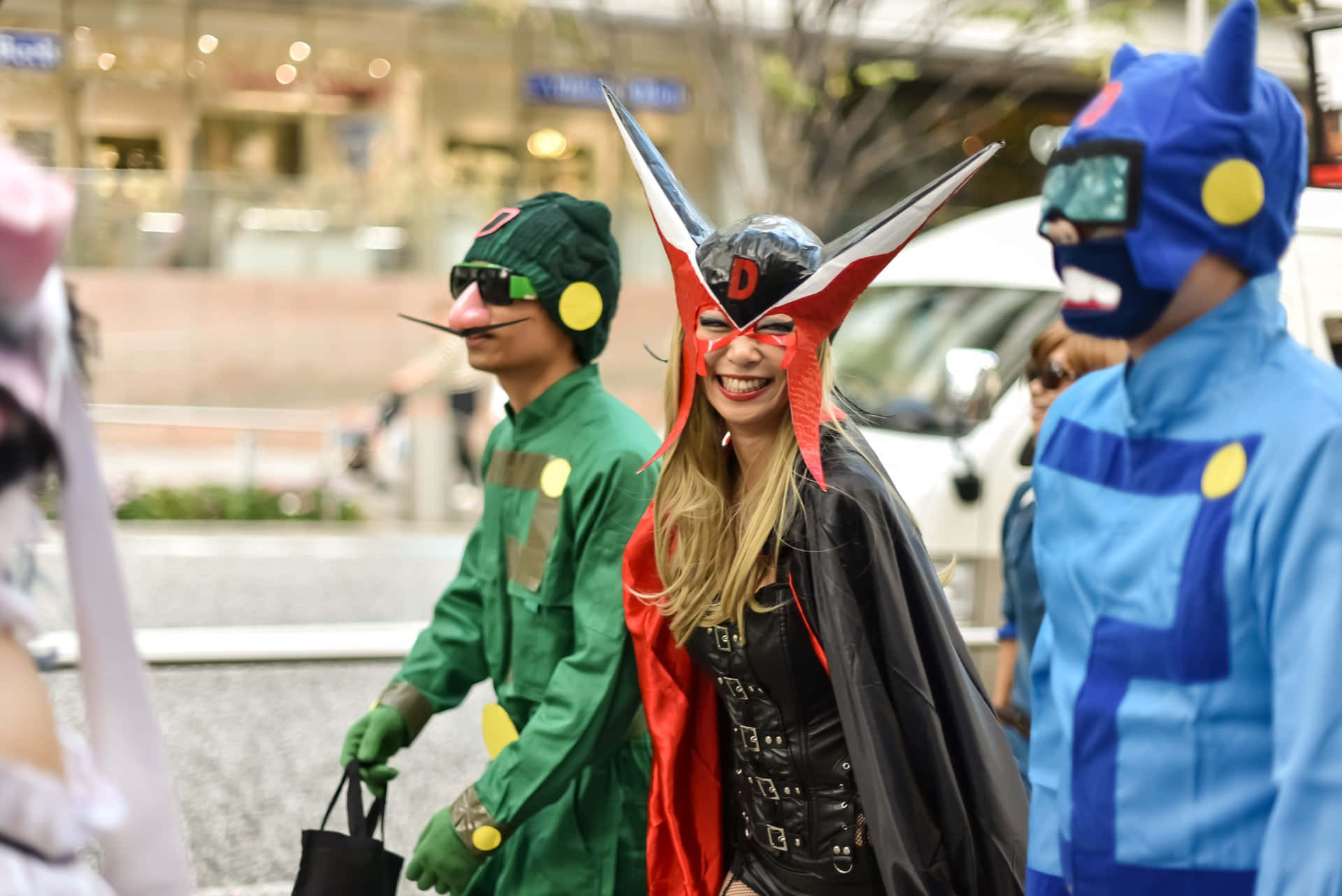 Halloween Parades 桌布 壁紙 — 三個穿著色彩繽紛動漫服裝的人，在模糊的商店櫥窗背景下，愉快地走在城市街道上。