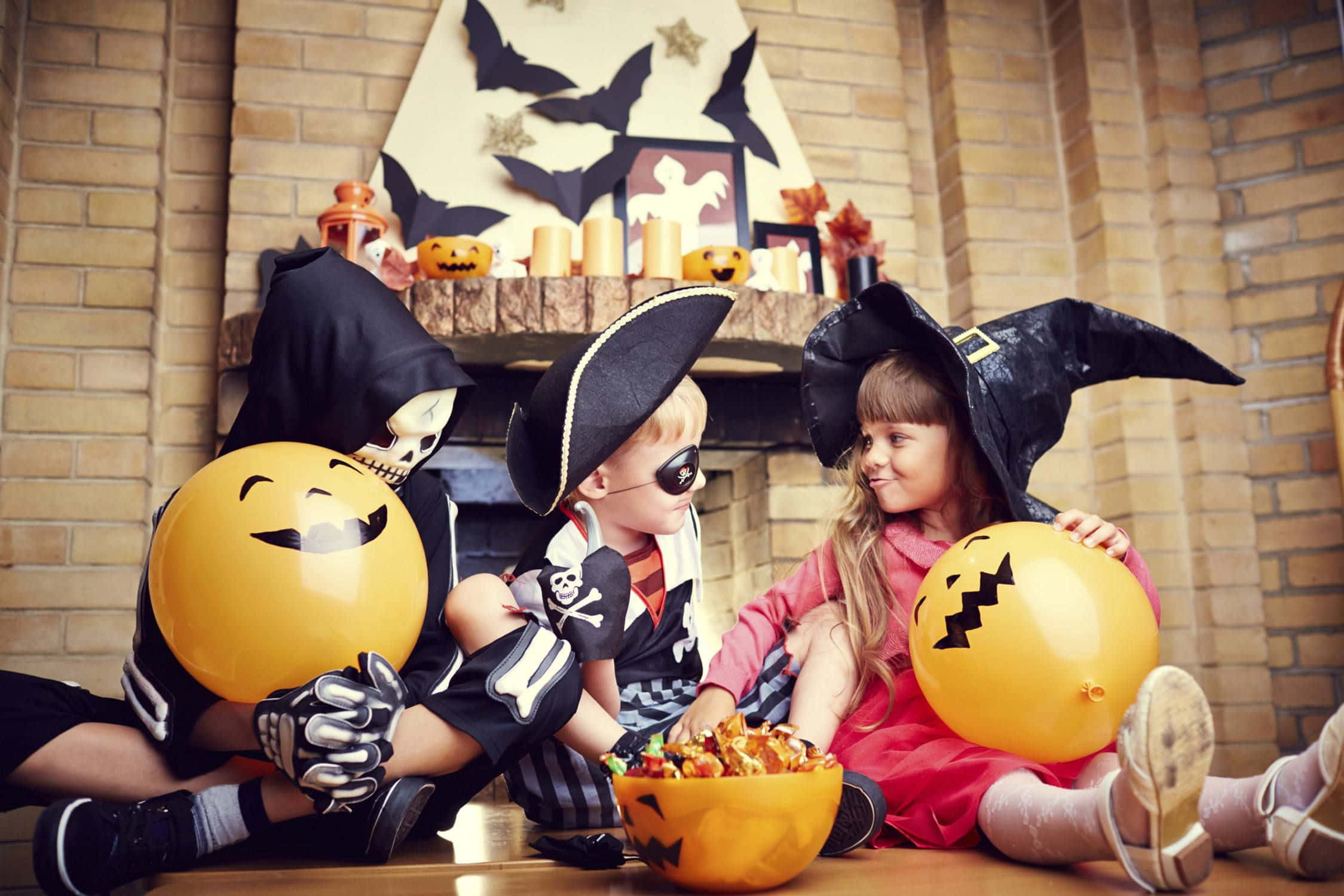 Halloween Costume 桌布 壁紙 — 三個穿著萬聖節服裝的小孩坐在木地板上，在裝飾過的磚砌壁爐旁拿著傑克南瓜燈氣球。