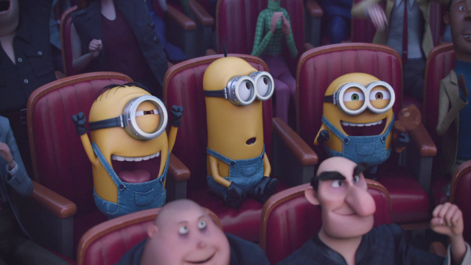 minions the rise of gru 桌布 壁紙 — 三個穿著藍色吊帶褲的黃色小小兵在紅色戲院座位上與周圍的人類一起看電影，開懷大笑。