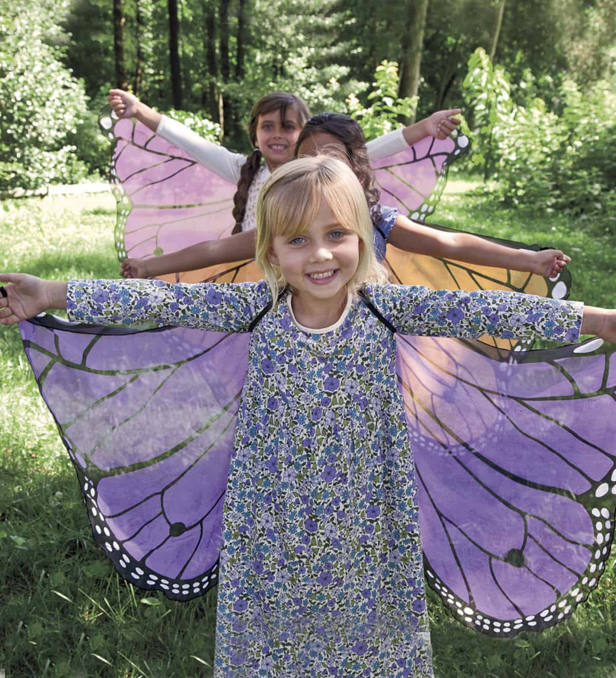 Butterfly Wing Dress 桌布 壁紙 — 三個笑容滿面的小孩在陽光照耀的森林中，戴著巨大的紫色蝴蝶翅膀，開心地張開雙臂擺姿勢。