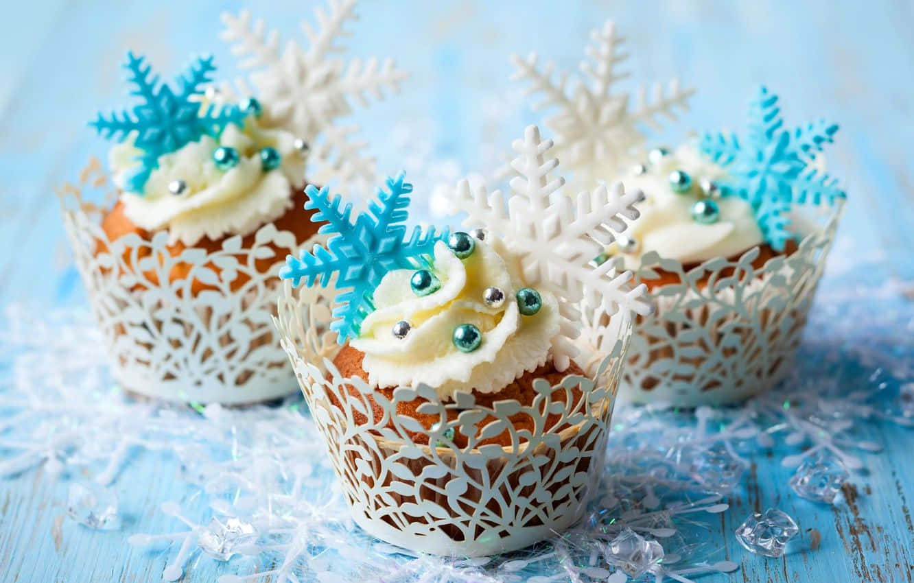 Winter Desserts 桌布 壁紙 — 三個節日紙杯蛋糕，上面有白色糖霜和藍色雪花裝飾，放在一個白雪覆蓋的藍色木質表面上。