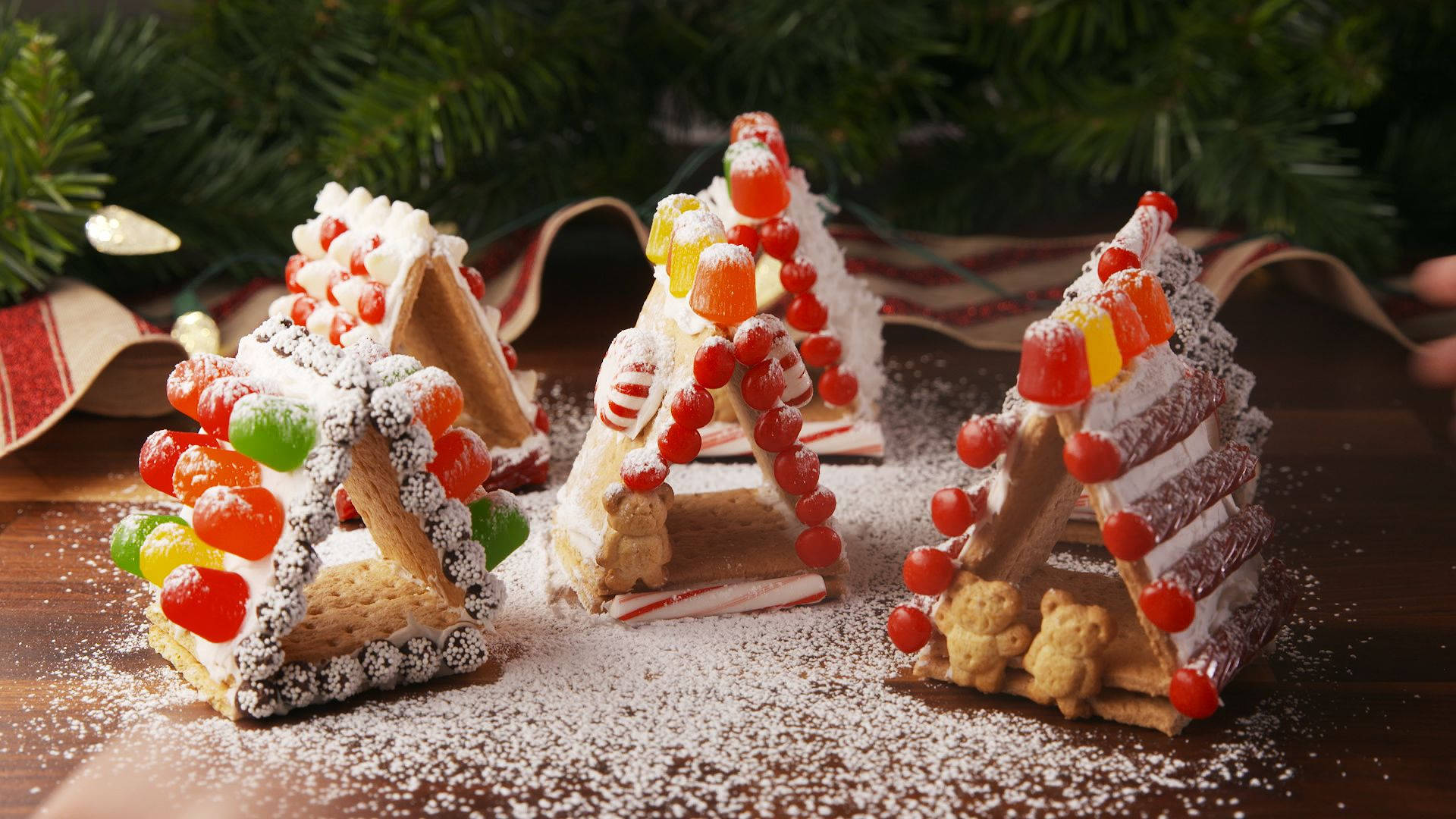 Gingerbread House 桌布 壁紙 — 三個節日薑餅帳篷，飾以彩色軟糖和糖粉，放在木桌上，旁邊有常青樹枝。