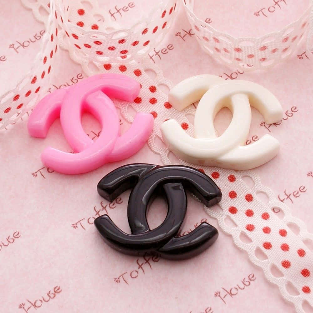 Chanel 桌布 壁紙 — 三個粉紅色、白色和黑色的光面 Chanel 標誌吊飾放在帶有紅色圓點和蕾絲邊的粉紅色布料上。