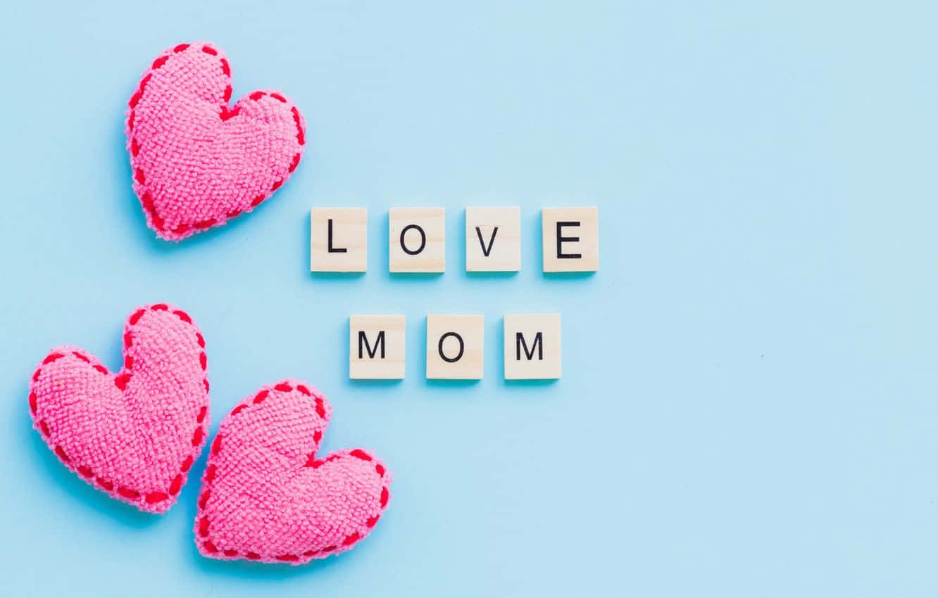愛心 桌布 壁紙 — 三個粉紅色布質心形和拼出「LOVE MOM」的木塊，排列在柔和的藍色背景上。