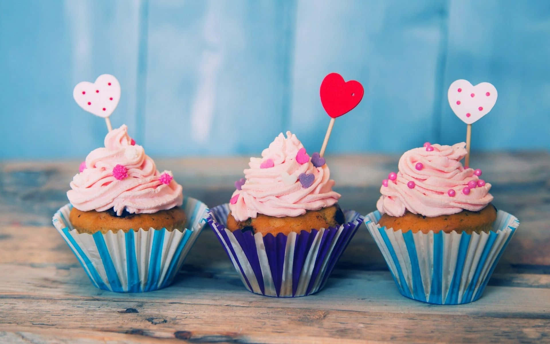 Cute Cupcake 桌布 壁紙 — 三個粉紅色糖霜杯子蛋糕，頂部有心形裝飾，放在質樸的木質表面上，背景是柔和的藍色。