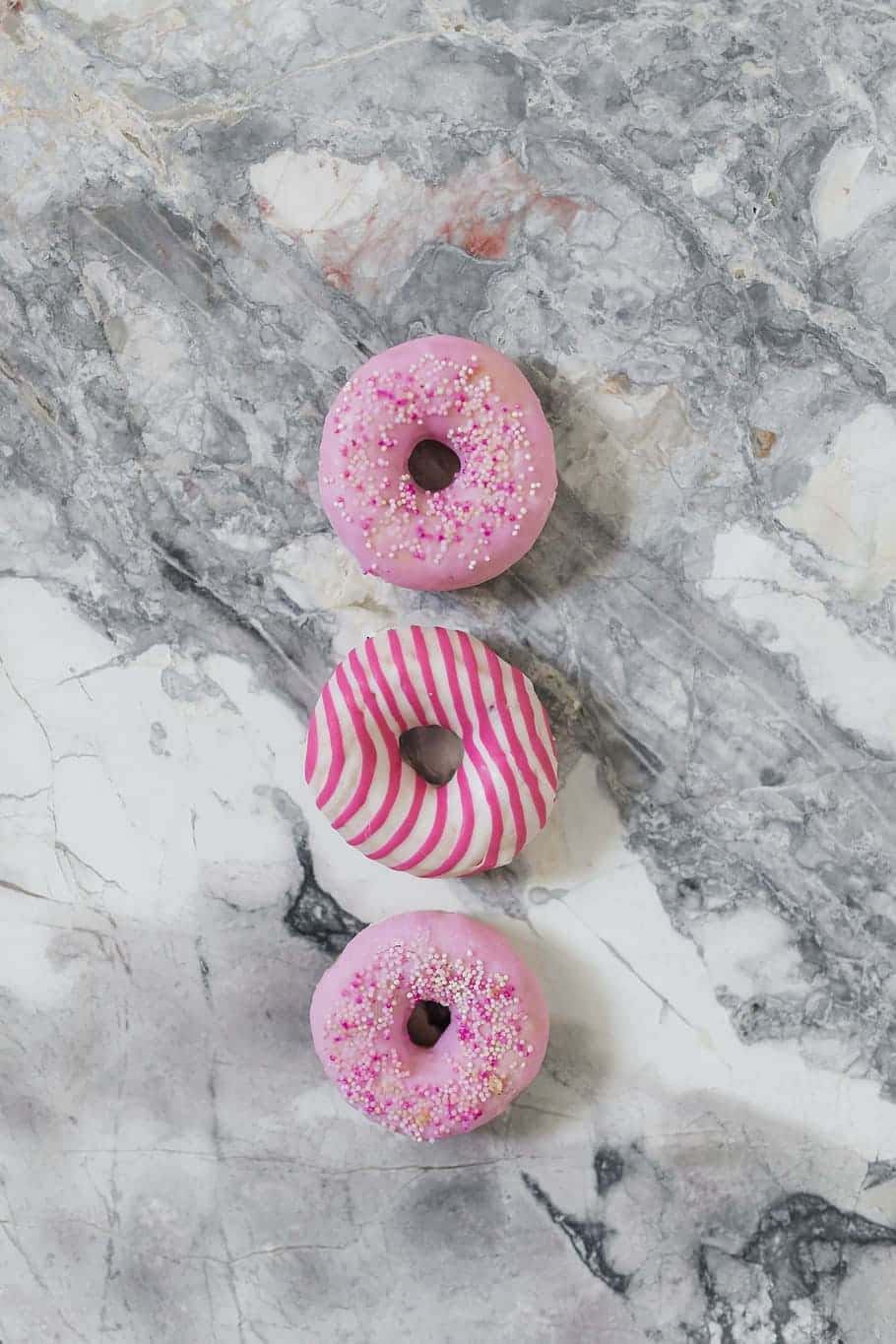 Cute Donut 桌布 壁紙 — 三個粉紅色糖霜甜甜圈，上面有彩色糖粒和條紋，垂直排列在灰色和白色的大理石表面上。