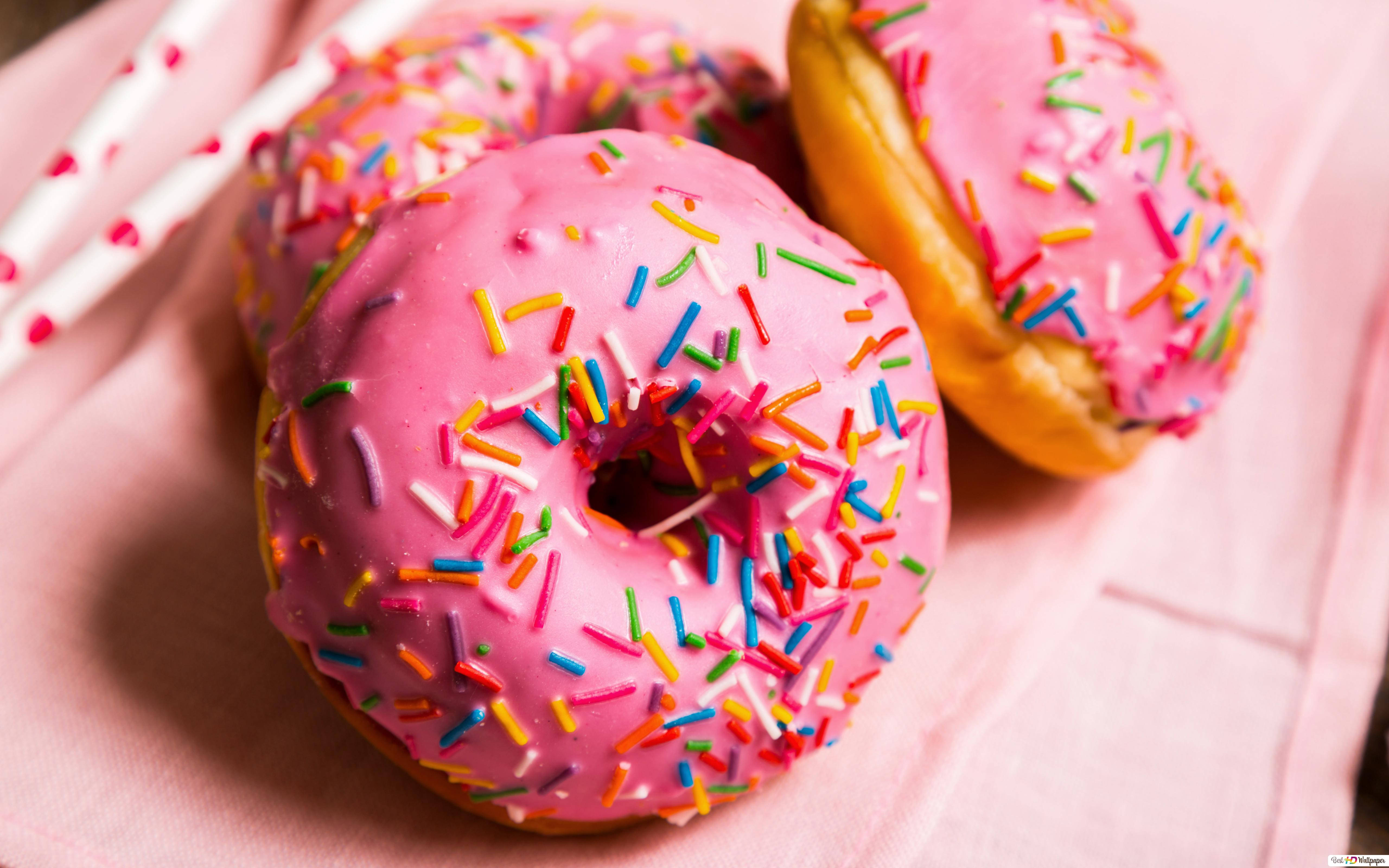 Donut 桌布 壁紙 — 三個粉紅色糖霜甜甜圈配上彩色糖粒，放在柔軟的粉紅色布上，旁邊有心形圖案的吸管。