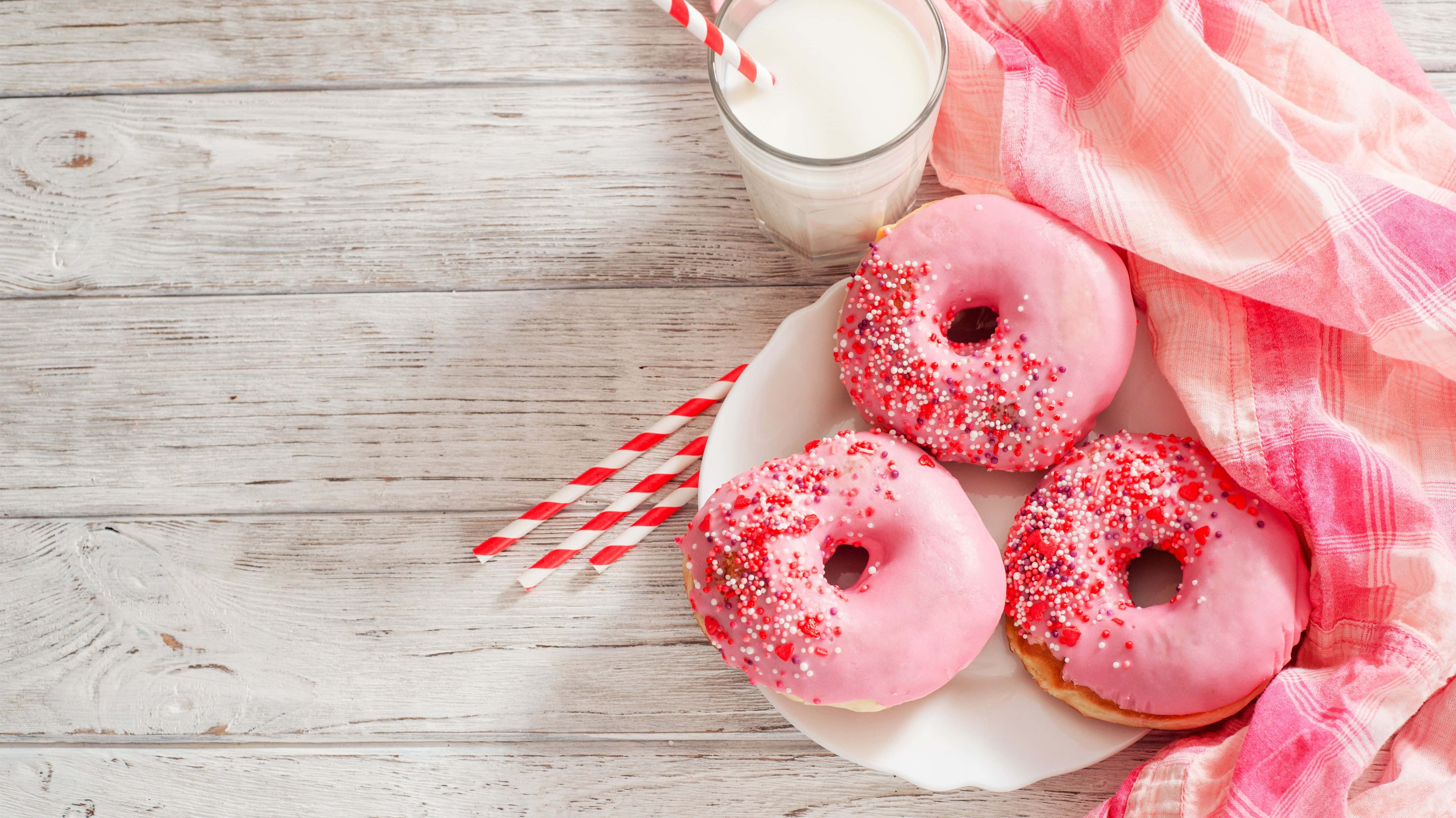 Cute Donut 桌布 壁紙 — 三個粉紅色糖霜甜甜圈配上紅色糖粒，放在白色盤子上，旁邊有一杯牛奶和一塊粉紅色布，置於風化的木桌上。