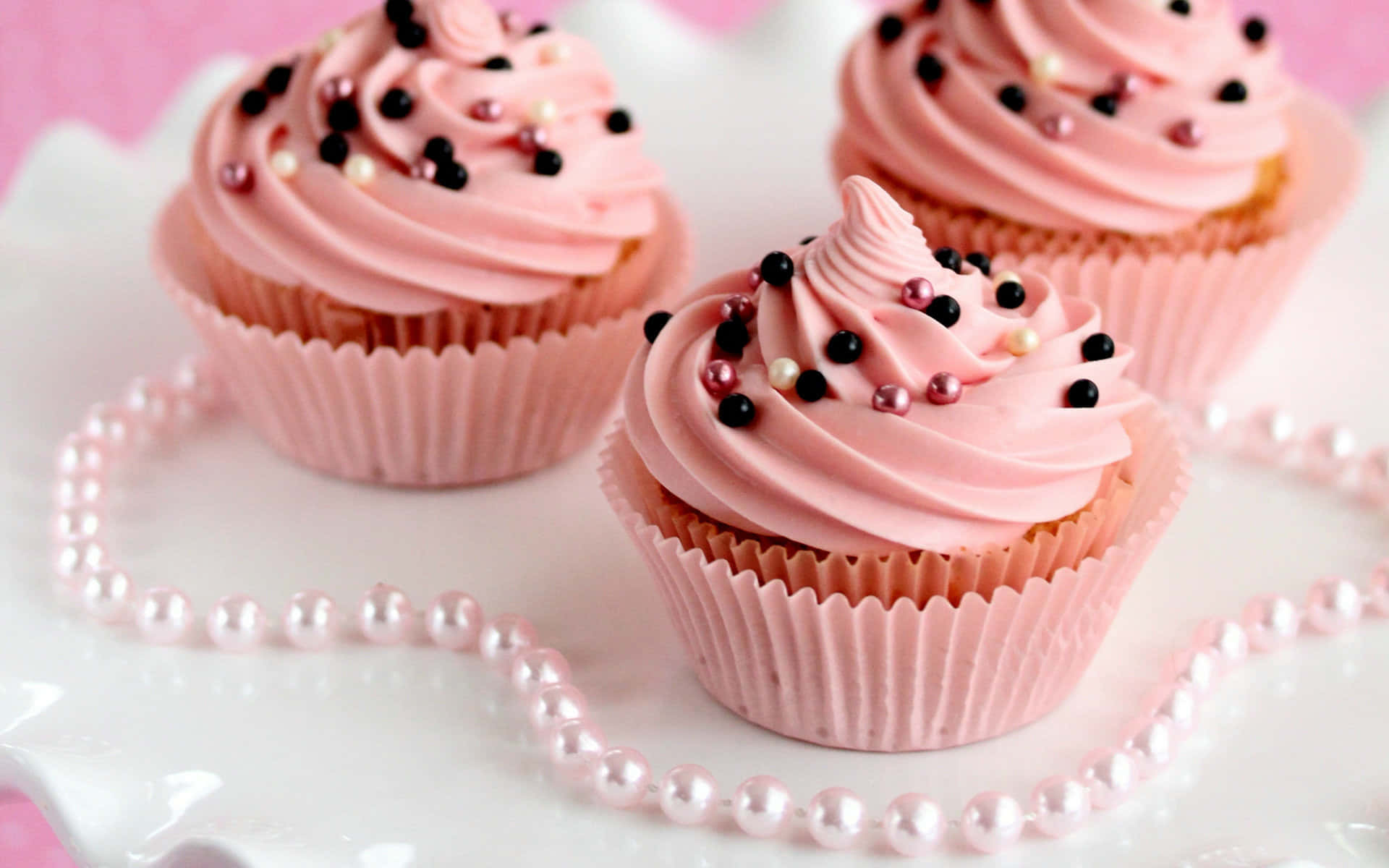 Cute Cupcake 桌布 壁紙 — 三個粉紅色糖霜紙杯蛋糕，上面有黑色和珍珠色糖粒，放在白色盤子上，珍珠項鍊形成心形。