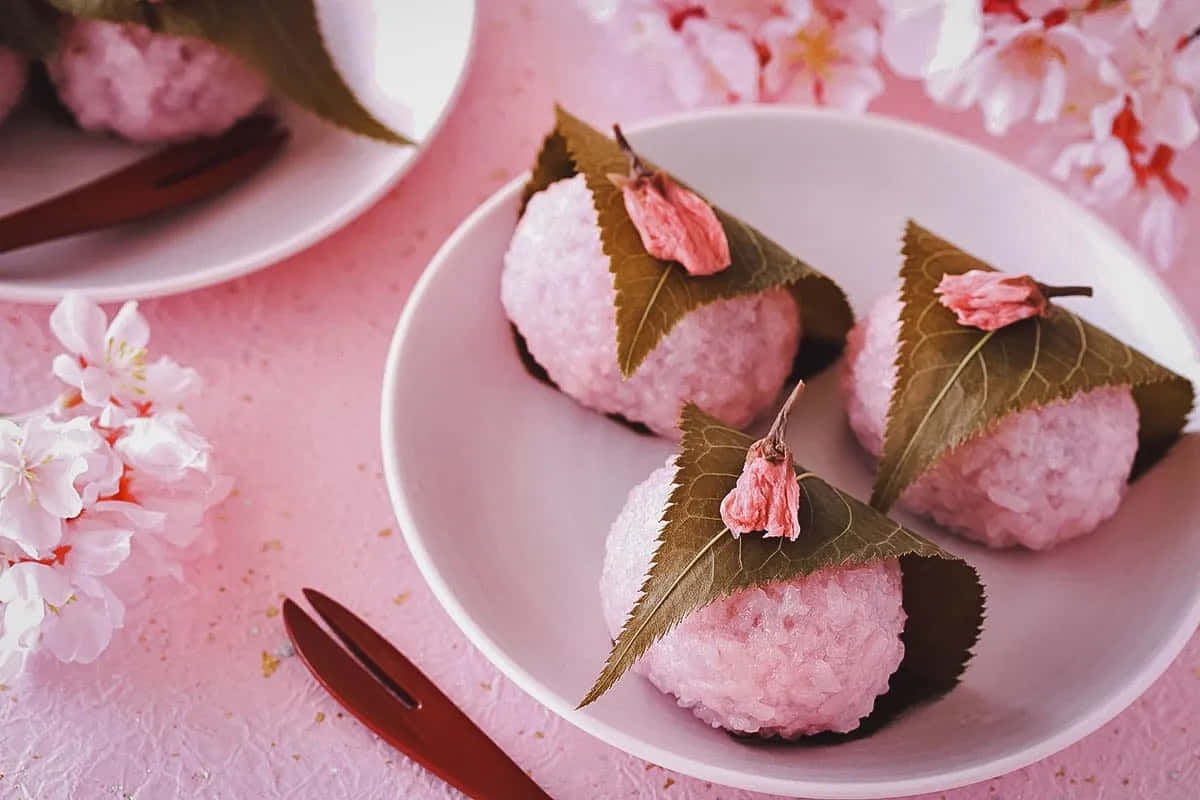 Cute Dessert 桌布 壁紙 — 三個粉紅麻糬球藏在綠葉中，上面有櫻花，置於柔軟的粉紅表面上。