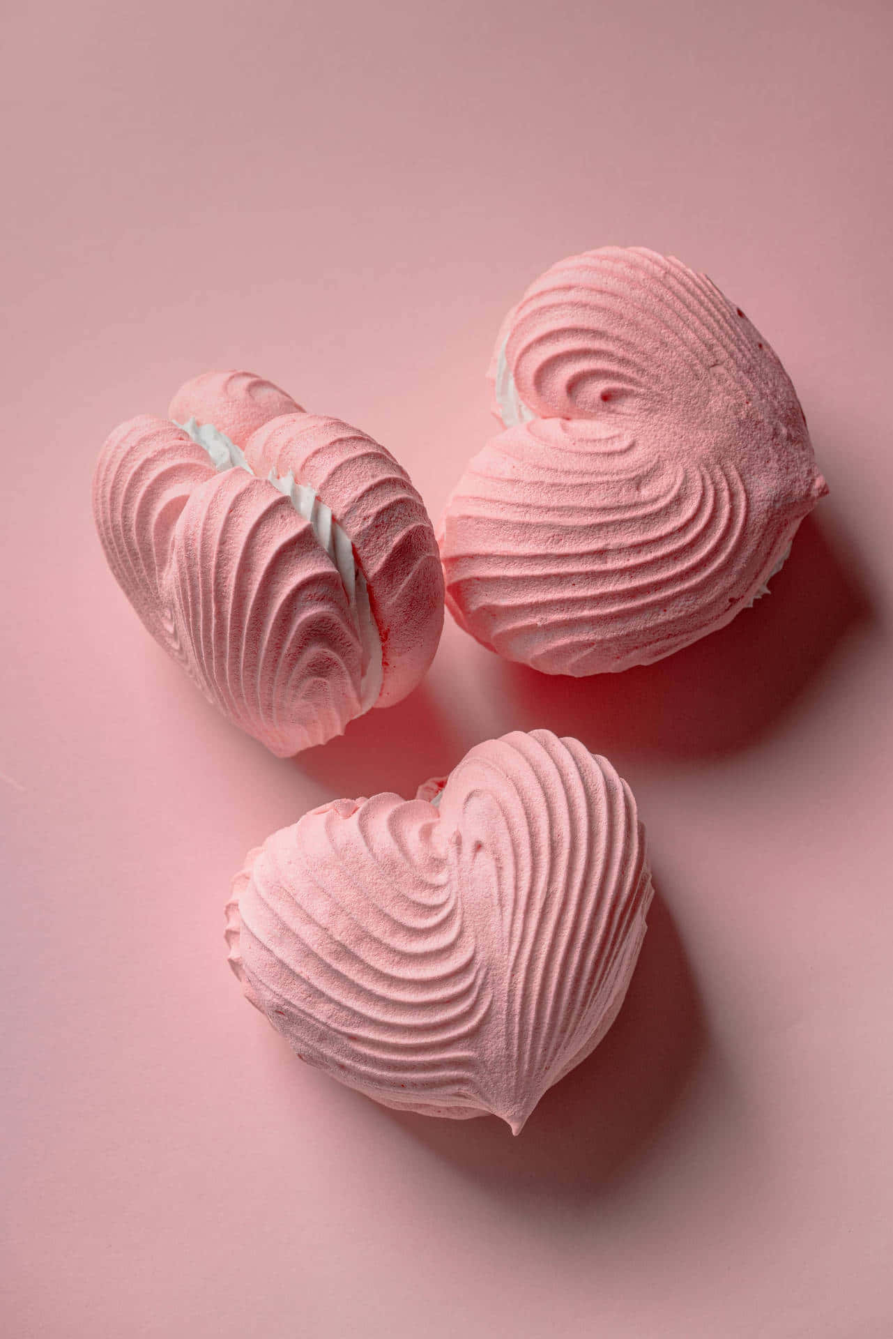 Light Pink iPhone 桌布 壁紙 — 三個精緻的粉紅色心形蛋白霜，帶有漩渦紋理，放置在柔軟的粉紅色表面上。