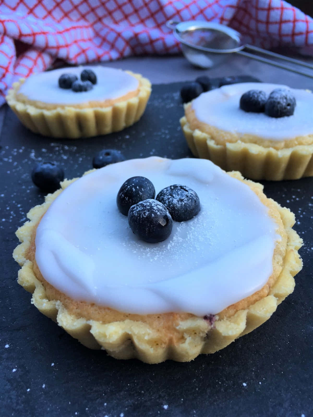 Blueberries Tart 桌布 壁紙 — 三個精緻的藍莓塔，帶有白色糖霜和糖粉，放在深色石板表面上，旁邊有一塊紅格紋布和篩子。