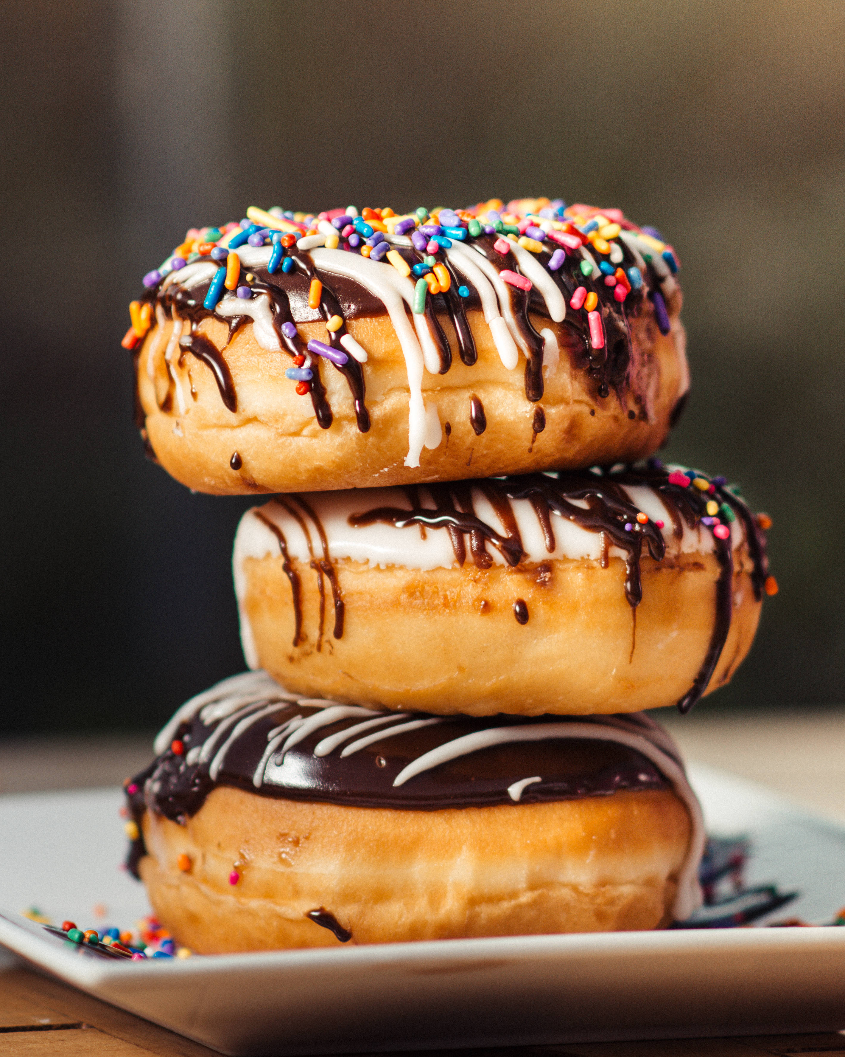 Dunkin Donuts 桌布 壁紙 — 三個糖霜甜甜圈高高疊起，上面有巧克力和白色糖霜滴落，並灑上彩色糖針，放在白色盤子上。