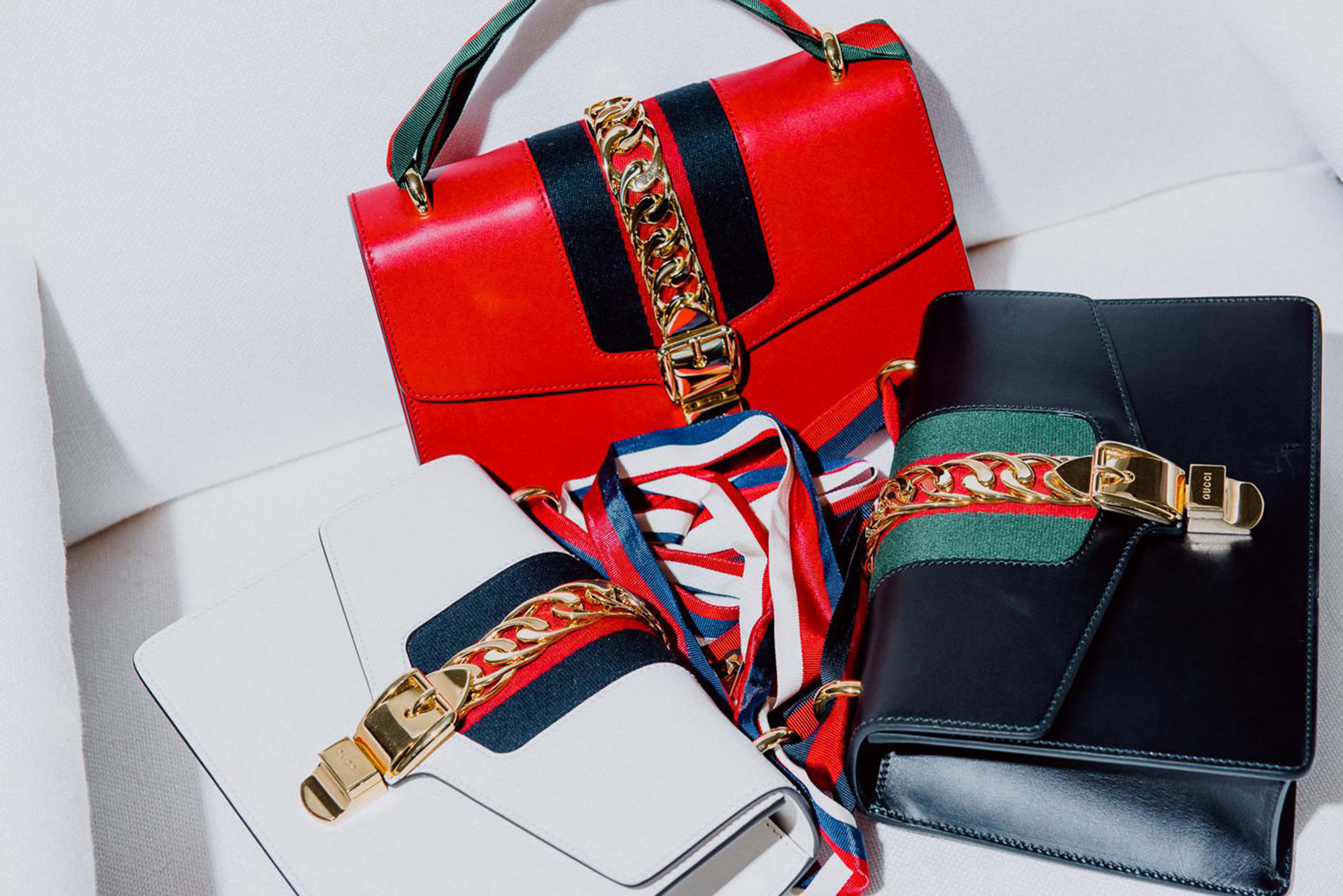 Gucci 桌布 壁紙 — 三個紅色、白色和黑色的奢華手提包，配有金色鏈條和條紋布料裝飾，擺放在白色表面上。