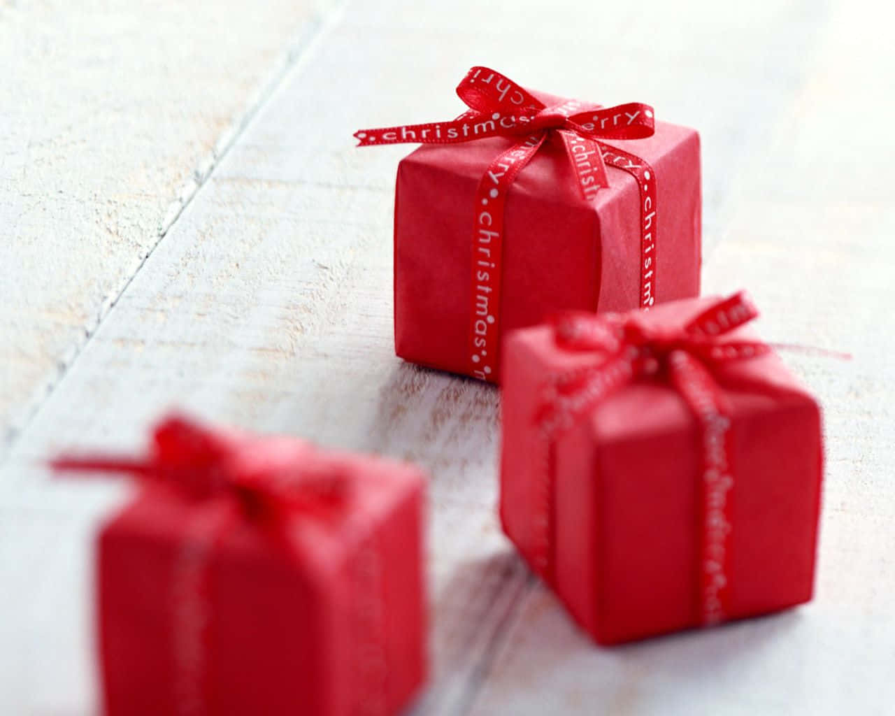 Gift 桌布 壁紙 — 三個紅色聖誕禮物盒，飾有節日絲帶，放在淺色木質表面上，一個清晰對焦，其他則柔和模糊。