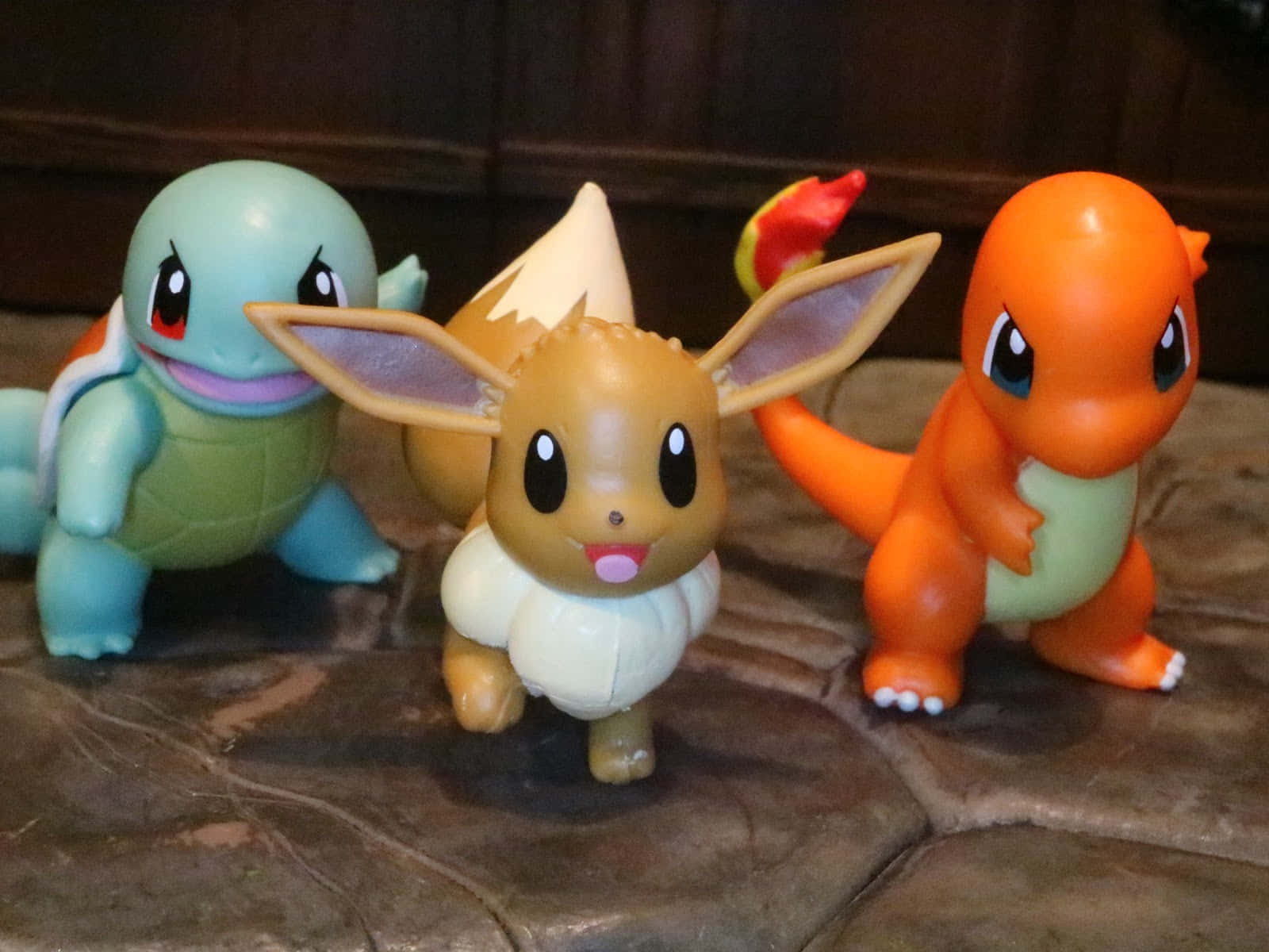 Pokemon Toys 桌布 壁紙 — 三個經典寶可夢公仔傑尼龜、伊布和小火龍在帶有紋理的棕色表面上一起擺姿勢。