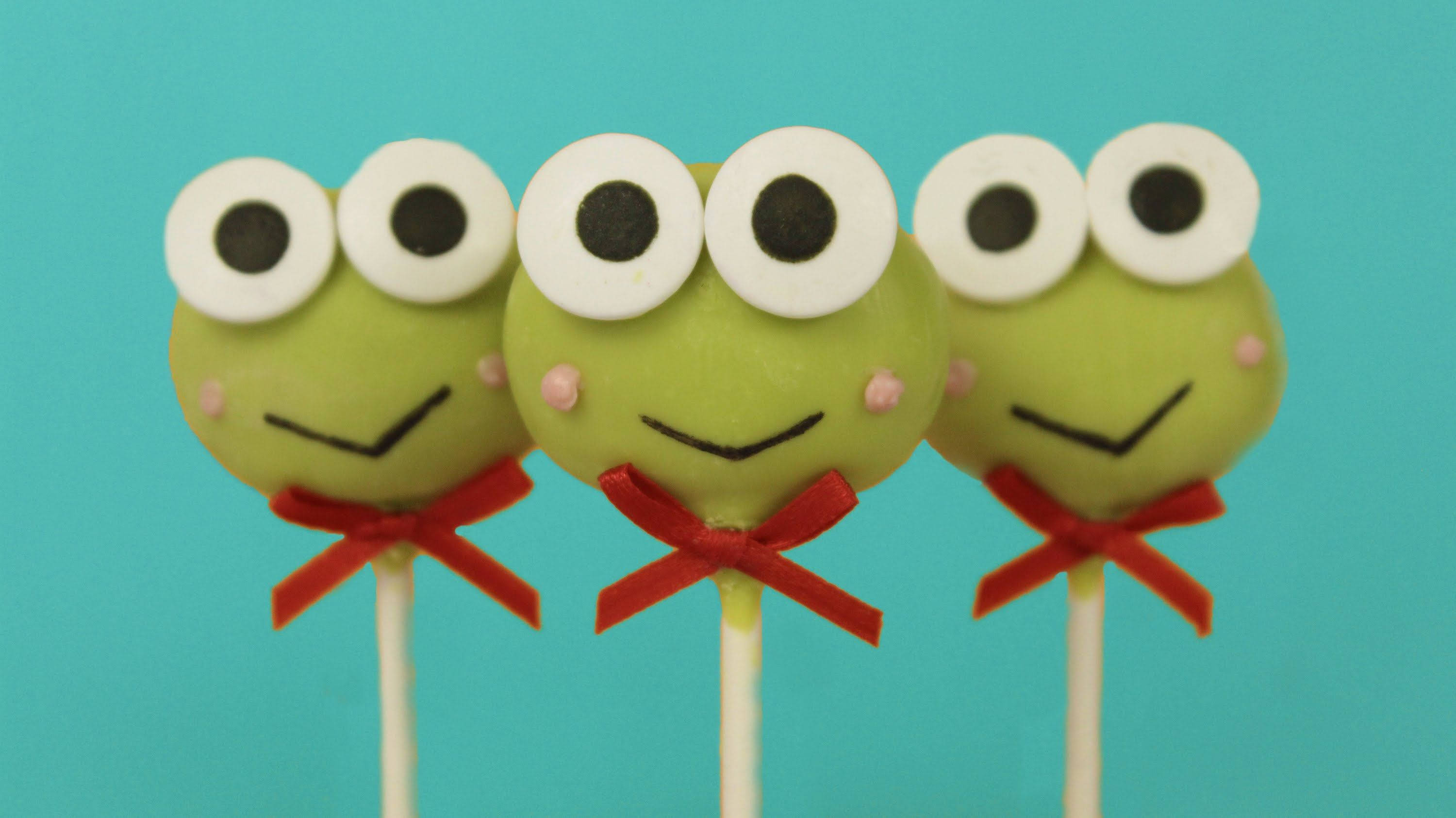 Keroppi 桌布 壁紙 — 三個綠色青蛙臉蛋糕棒棒糖，有大眼睛和紅色蝴蝶結，站在藍綠色背景前。